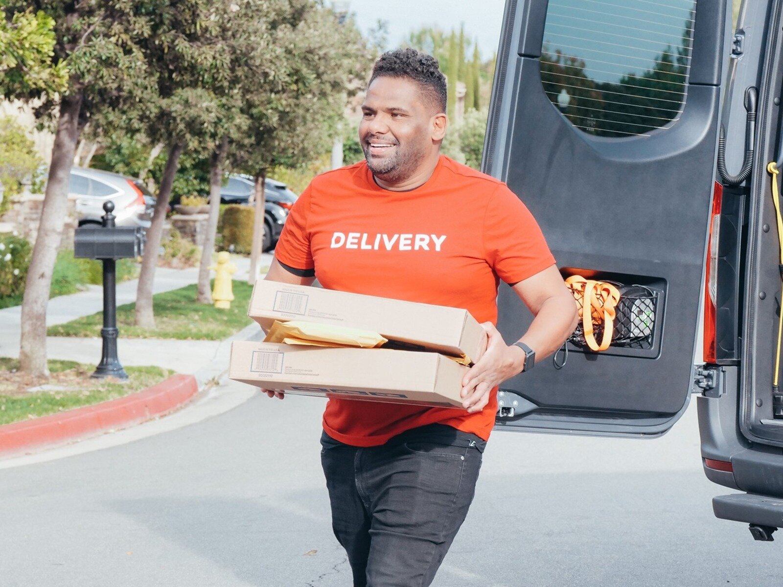 Zdjęcie przedstawia młodego mężczyznę, który trzyma w dłoniach dwie płaskie paczki w tekturowym opakowaniu. W tle widoczne są uchylone tylne drzwi samochodu dostawczego. Mężczyzna nosi pomarańczową koszulkę z napisem w języku angielski: DELIVERY. 