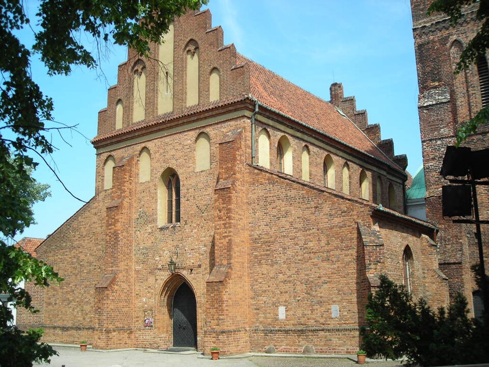Fotografia przedstawia budynek zbudowanych z cegły. Główna część ma kształt prostokąta, od której po obu stronach odchodzą niższe elementy o spadzistych dachach. Zarówno okna, jak i wejście zwieńczone są ostrymi łukami. Po prawej stronie do budynku przylega wieża.