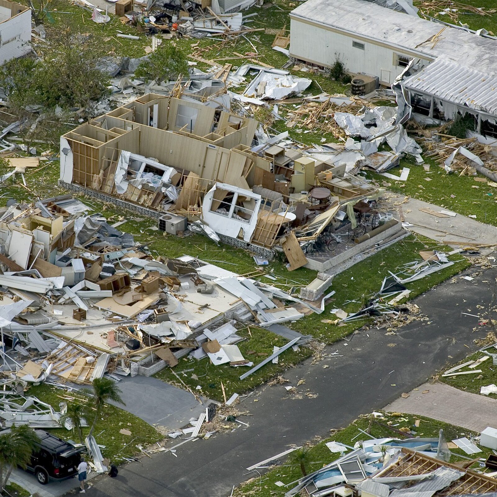Zdjęcie przedstawia zniszczenia spowodowane przez huragan. Osiedle drewnianych domów doszczętnie zniszczone. Większość domów nie ma dachów ani ścian. Cała ziemia pokryta fragmentami zniszczonych domów.