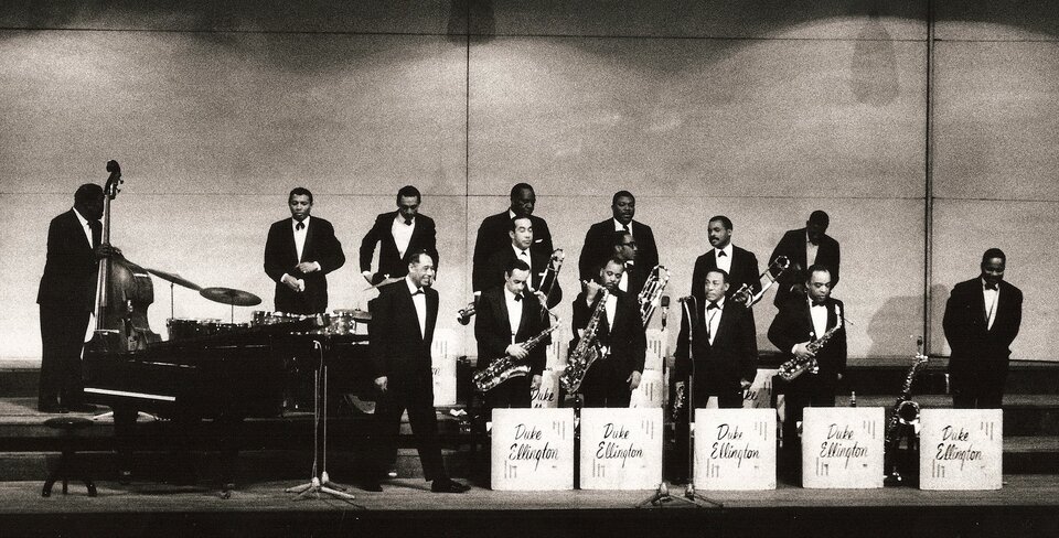 Na czarno‑białym zdjęciu big band Duke'a Ellingtona. Na zdjęciu trzynastu mężczyzn. Widać fortepian, perkusję i kontrabas. Mężczyźni, grający na instrumentach dętych, stoją w dwóch rzędach. W pierwszym rzędzie saksofoniści. W drugi grający na instrumentach dętych blaszanych.