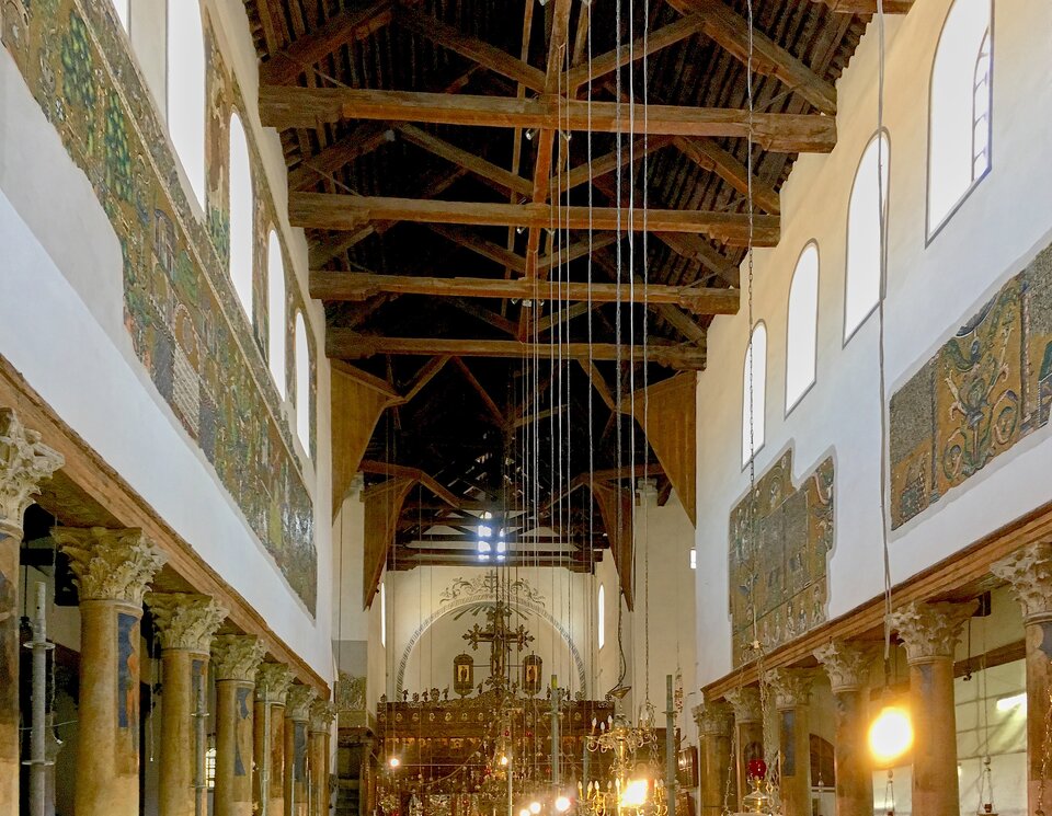 Wnętrze bazyliki z długą nawą główną: rzędy kamiennych kolumn z bogato rzeźbionymi kapitelami podtrzymują arkady, nad którymi widoczne są fragmenty starożytnych mozaik. Drewniany, otwarty strop z belkami biegnie wzdłuż całej nawy, a w głębi znajduje się bogato zdobiony ikonostas i ołtarz, oświetlone wiszącymi lampami i świecznikami.