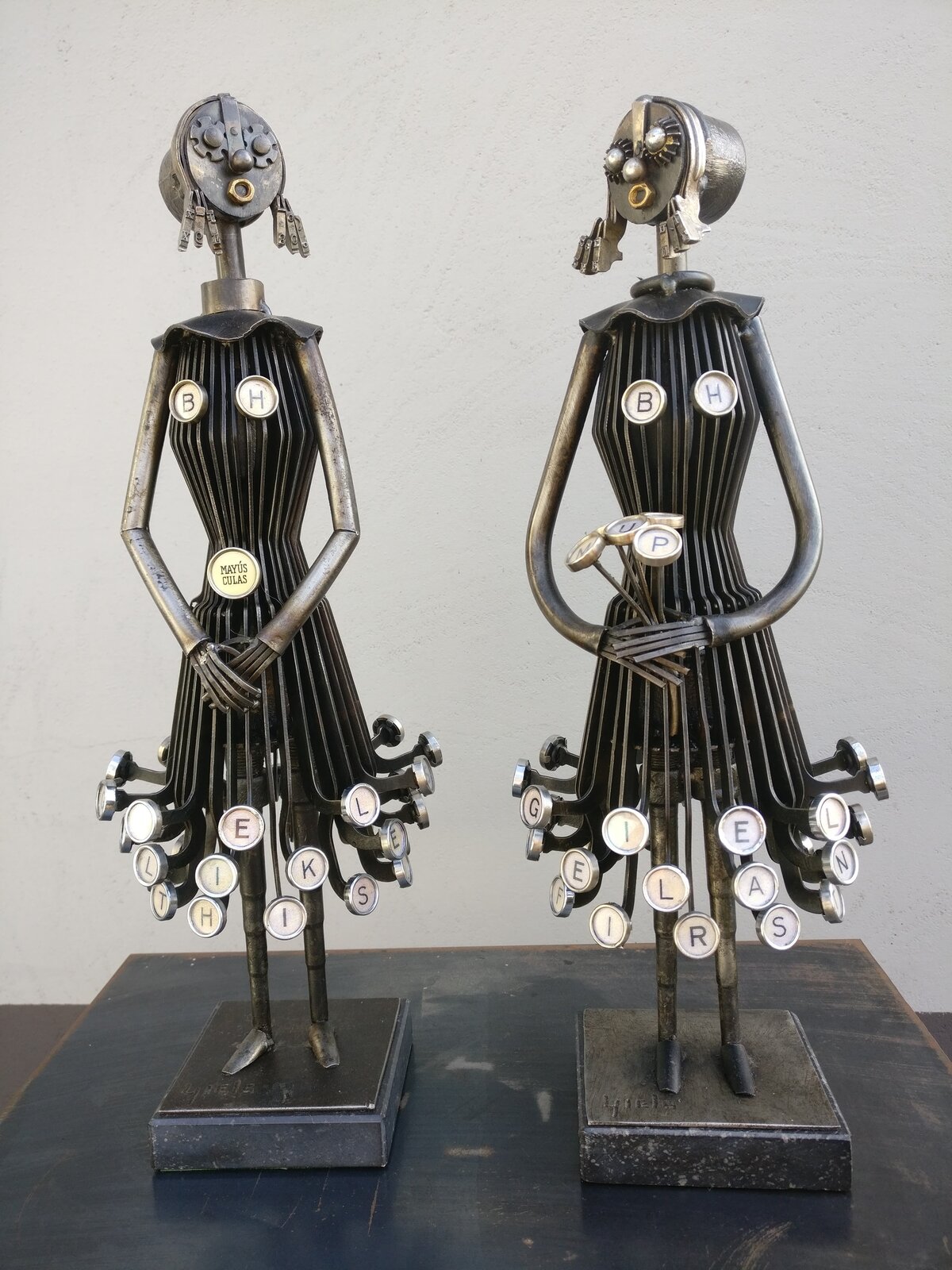 Ilustracja przedstawia asamblaż Ludo Giels skonstruowany z maszyny pisarskiej. Instalacja to dwie postacie kobiet wykonane z metalu. Stoją obok siebie trzymając ręce złożone. Jedna z nich trzyma bukiet kwiatów wykonany z liter z maszyny do pisania. Litery są również u dołu sukni obu kobiet.