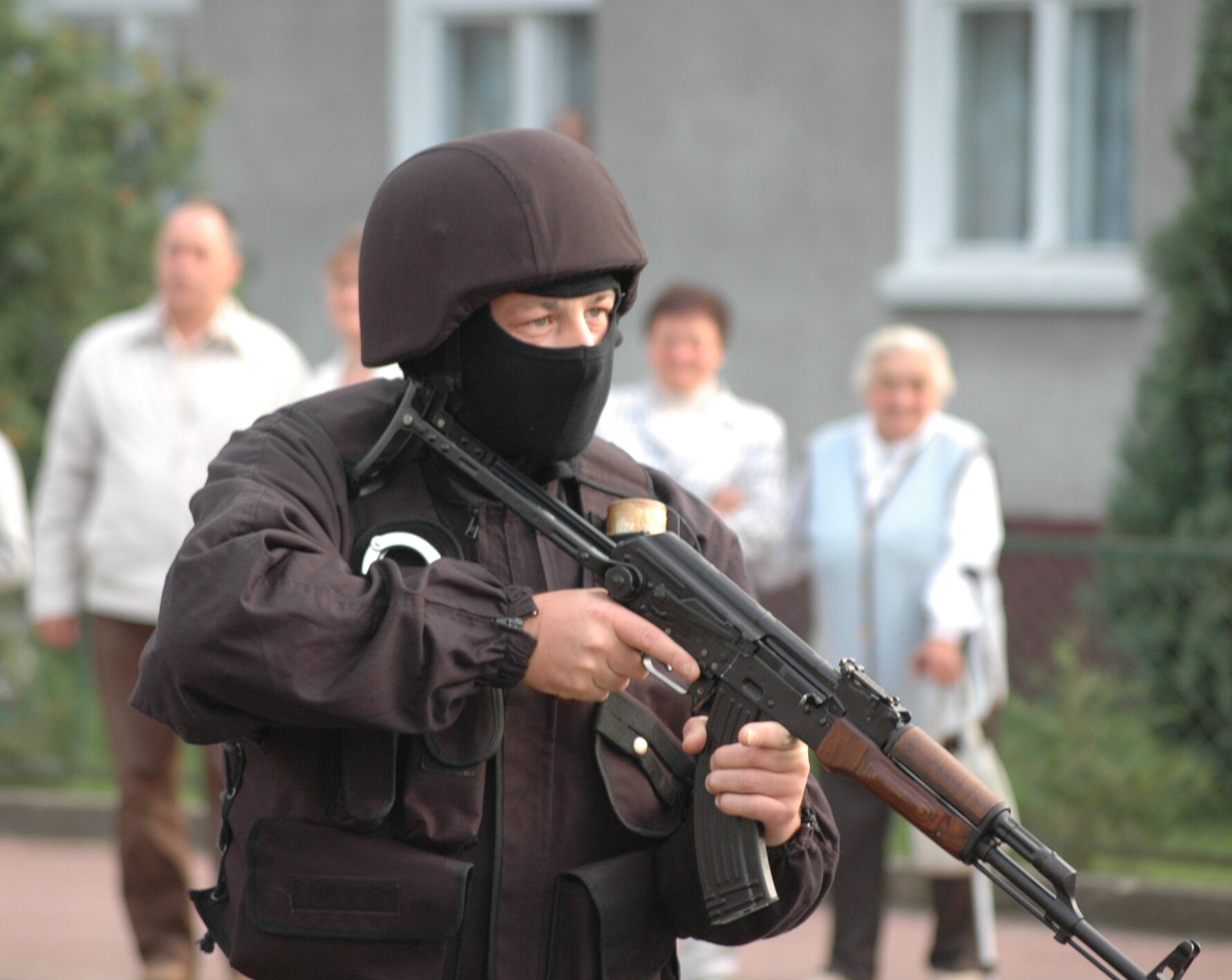 Zdjęcie przedstawia mężczyznę w czarnym mundurze. Na głowie ma hełm. Na twarzy czarną kominiarkę. Trzyma karabin. W tle stoi kilka starszych osób i przygląda się mężczyźnie.     
