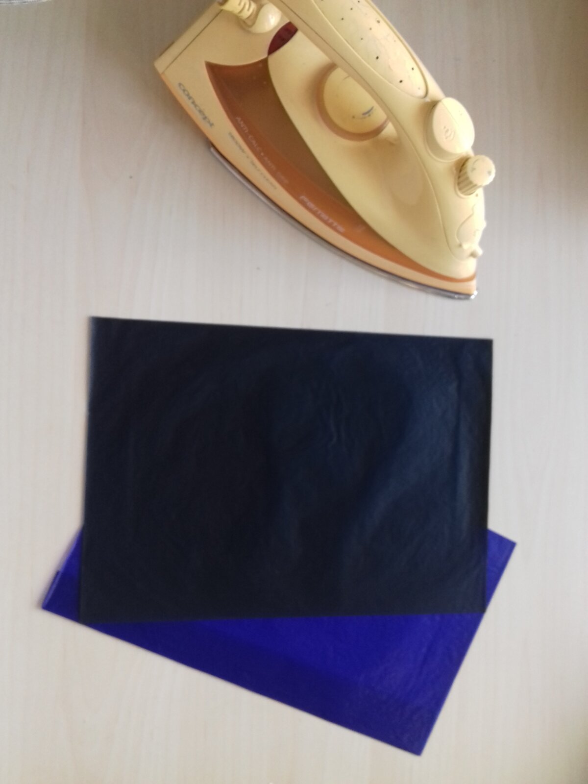 Ilustracja przedstawia blat stołu, na którym leżą dwie kalki maszynowe w kolorach czarnym i niebieskim. Obok nich postawione jest żółte żelazko.