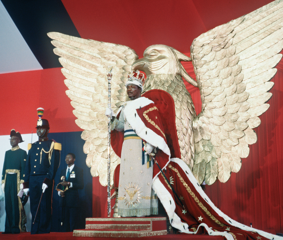 Fotografia przedstawia czarnoskórego mężczyznę w królewskich szatach. Mężczyzna stoi. Na głowie ma koronę zdobioną drogocennymi kamieniami, w jednym ręku trzyma berło, w drugiej szablę przy boku. Za władcą stoi złoty posąg ptaka z rozpostartymi skrzydłami. Po lewej stronie widać czarnoskórego żołnierza z szablą przy boku i z hełmem na głowie.