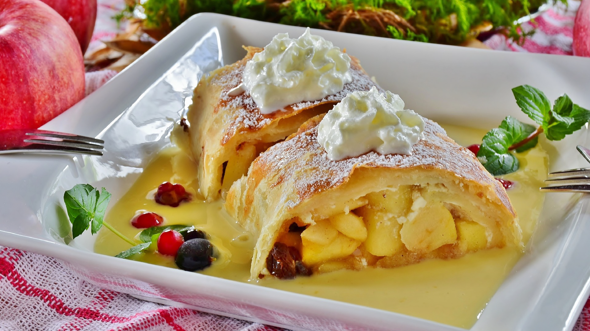 Na zdjęciu pokazany jest Strudel z bitą śmietaną, wypełniony jabłkami i posypany cukrem pudrem. Strudel podany jest na głębokim talerzu.