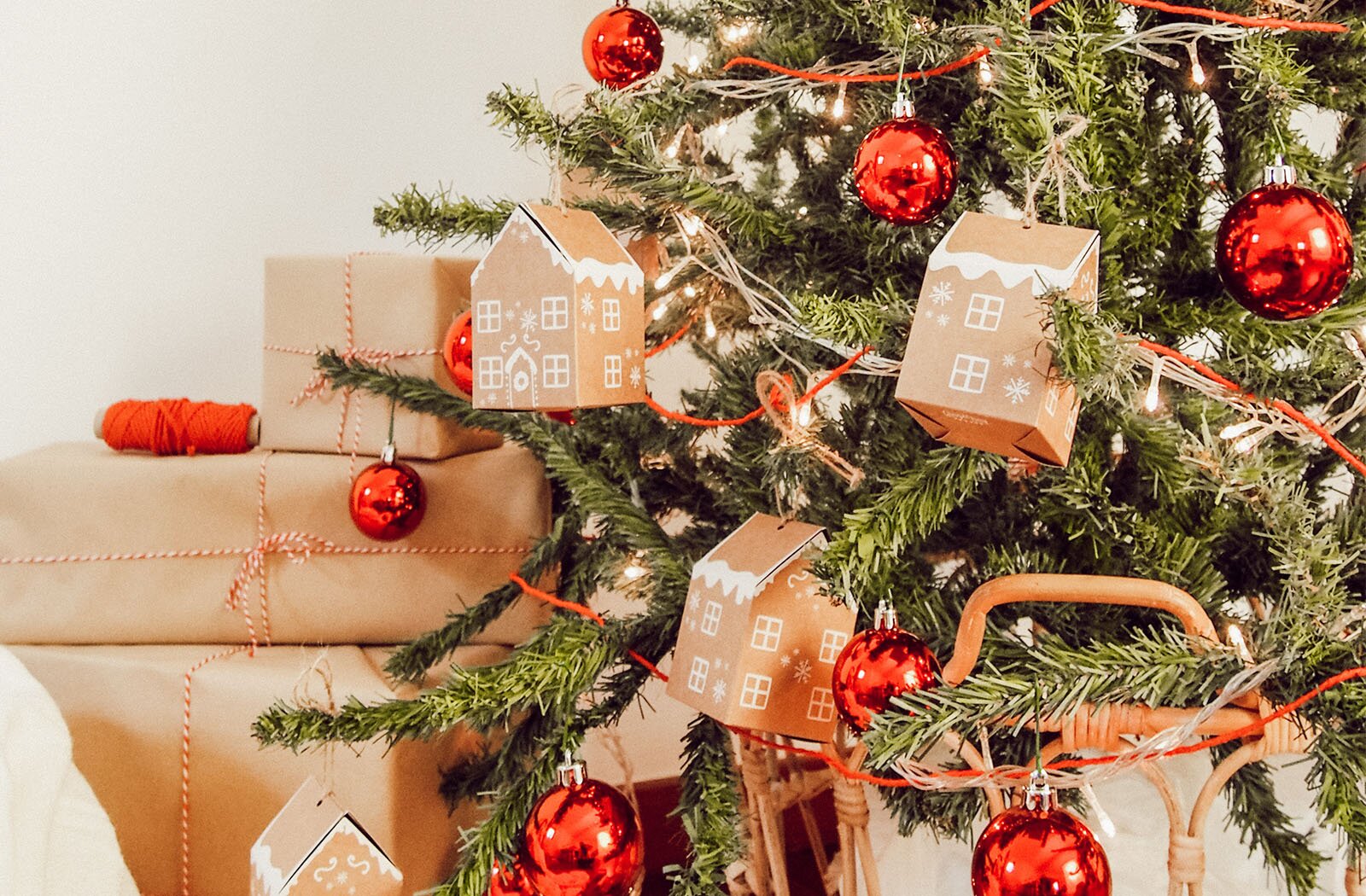 Zdjęcie przedstawia fragment choinki z prezentami. Po prawej stronie widoczny jest świerk, ustawiony na wiklinowym stojaku i udekorowany czerwonymi bombkami, światełkami oraz kartonowymi domkami ze świątecznym ornamentem. Na lewo widoczne są trzy, ustawione na sobie pudełka prezentowe, w różnym rozmiarze, owinięte szarym papierem i przewiązane biało‑czerwonym sznurkiem.