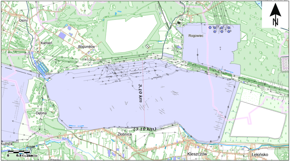 Mapa przedstawia okolice odkrywki węgla brunatnego Bełchatów. W centralnej części widoczny jest rozległy obszar wyrobiska zaznaczony kolorem jasnofioletowym. Na mapie naniesiono liczne strzałki pokazujące kierunki ruchu mas ziemnych oraz przerywaną czarną linię z oznaczeniem odległości 3,10 km, przebiegającą przez środkową część wyrobiska. Wokół odkrywki znajdują się tereny leśne, pola uprawne, drogi oraz miejscowości: Rogowiec, Bogumiłów, Kamień, Osiny, Dębina, Złobnica, Kleszczów i Łękińsko. W prawym górnym rogu umieszczono strzałkę północy, a w lewym dolnym rogu podziałkę mapy.
