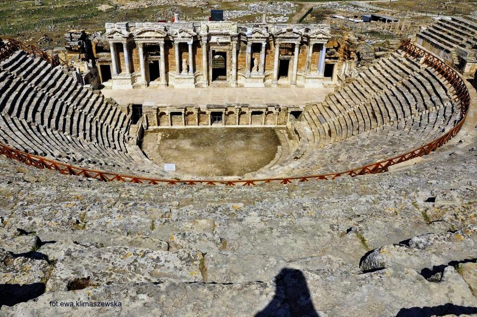 Zdjęcie przedstawia starożytną budowlę z bloków kamiennych. To amfiteatr w Hierapolis z III wieku n.e. Podobny jest do widowni stadionu, gdzie miejsca dla widzów pną się w górę na przeszło dwadzieścia poziomów. Amfiteatr zbudowany jest na planie połowy koła. W dole znajduje się szeroka scena w kształcie prostokąta, którą zamyka ściana, przypominająca dekoracyjną budowlę, z kolumnami i wejściami dla aktorów. Amfiteatry starożytne miały doskonałą akustykę, podobno głosy ze sceny słychać było nawet w ostatnim rzędzie.