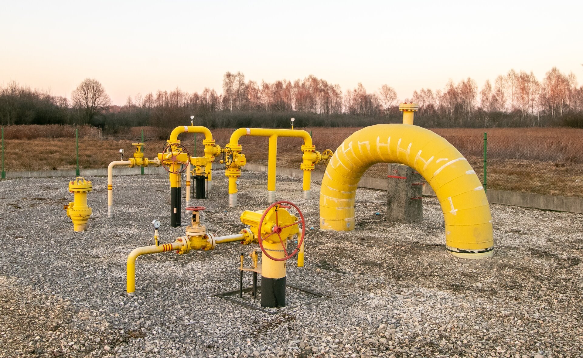 Na zdjęciu przedstawiono układ rurociągów z zasuwami odcinającymi na gazociągu przesyłowym i obiegach technologicznych. Widoczna struktura składa się z rur wychodzących w ziemi. W oddali, po prawej stronie widać  układ dwóch rur wychodzących z ziemi które są połączone ze sobą. Układ tworzy kształt obróconej litery U. Rura łączą dwie wychodzące z ziemi rury jest połączona przy użyciu połączenia kołnierzowego. Poniżej połączenia znajdują się zawory z kurkami o kształcie dużych czerwonych okręgów. Po środku rury widoczny jest okrągły wskaźnik ze wskazówką. Na zdjęciu znajdują się dwa takie układy. Układy są połączone przy użyciu elementu z dużym okrągłym kurkiem o kształcie koła.Po prawej stronie rysunku widać żółtą rurę o dużej średnicy wychodząc i wchodzącą pod ziemie. Rura ma kształt odwróconej litery U. Po lewej stronie rysunku znajduje się pionowa rura wychodząca z ziemi. Na końcu rury znajduje się duży czerwony kurek o kształcie okręgu. Cały układ jest zabezpieczony ogrodzeniem.