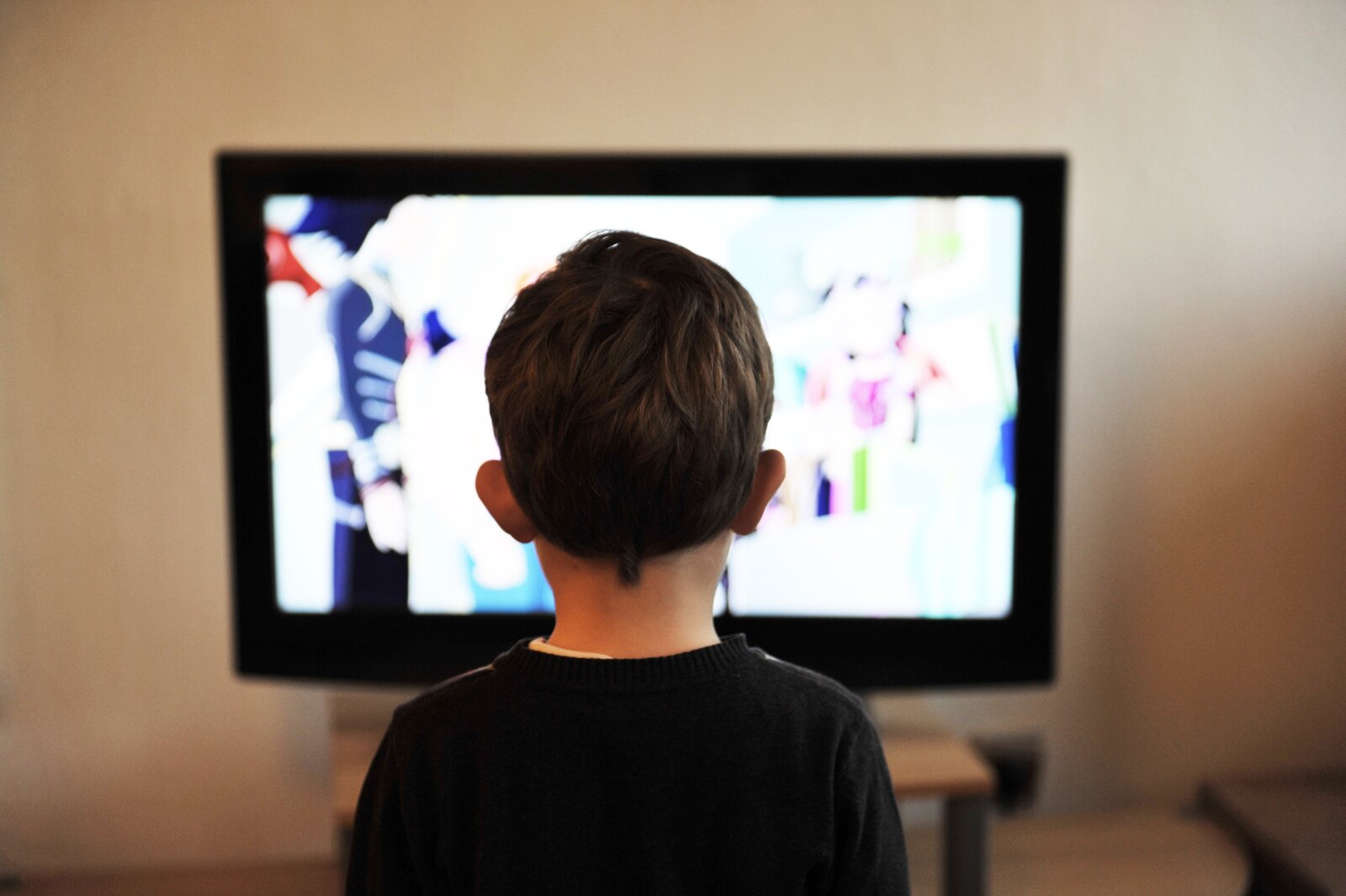 Fotografia przedstawia chłopca, który siedzi tyłem przed ekranem włączonego telewizora. Ma krótkie włosy i ciemną bluzkę. Na ekranie wyświetla się rozmyty jasny obraz. Telewizor stoi na tle białej ściany.
