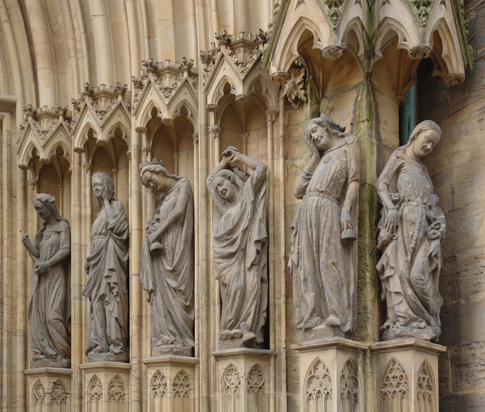Fotografia przedstawia dekorację rzeźbiarską portalu katedry w Erfurcie w Niemczech. Na zdjęciu widocznych jest sześć stojących postaci kobiecych. Stoją na zdobionych postumentach w kształcie prostokąta, są wsunięte w głąb muru. Nad postaciami znajdują się ozdobne daszki. Postaci są z jasnego kamienia, mają długie szaty, są wygięte w różne strony. Mają długie włosy, niektóre z nich podpierają głowę rękami. 
