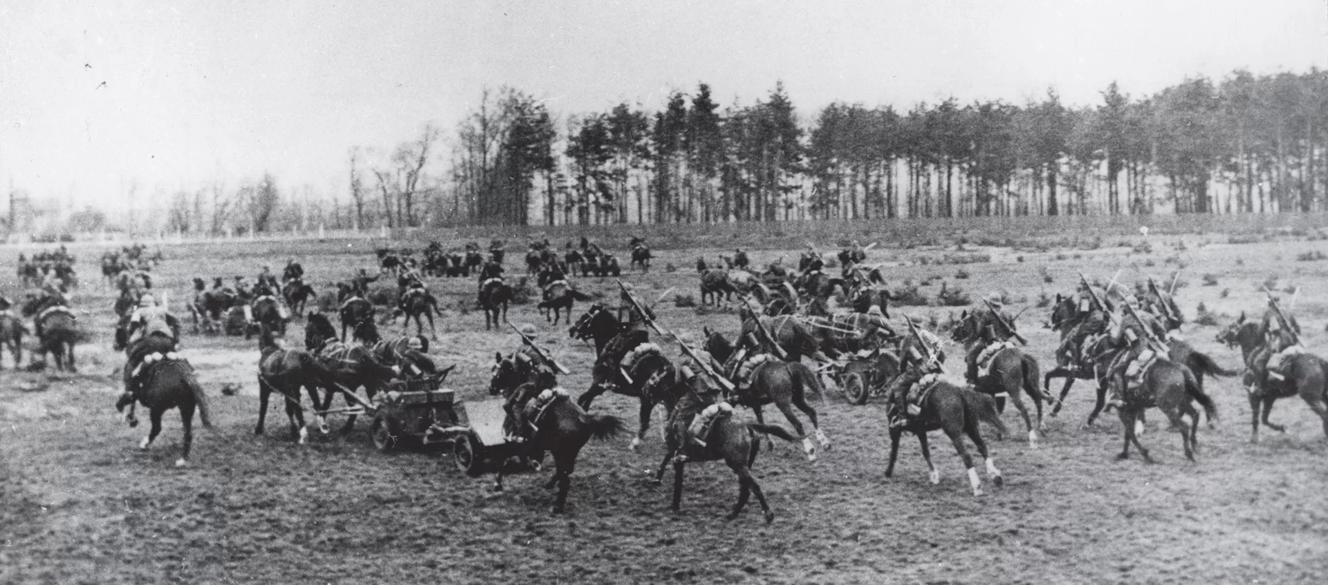 Zdjęcie biało‑czarne przedstawiające pluton artylerii przeciwpancernej 17 Pułku Ułanów Wielkopolskich podczas bitwy nad Bzurą we wrześniu 1939 r. Ułani jadą na czarnych koniach. Na drugim planie znajduje się niewielki las.