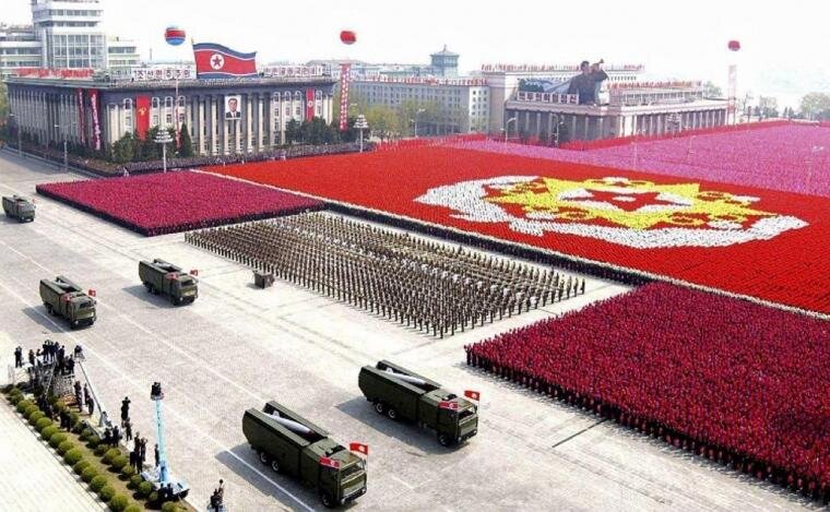Zdjęcie przedstawia rozległy plac, na którym odbywa się wojskowa defilada. Ulicą podążają dwójkami wozy z wyrzutniami rakiet. Wzdłuż drogi ich przejazdu znajdują się żołnierze w szyku oraz stoi tłum ludzi, tworząc ogromną czerwoną płaszczyznę. Pośrodku tej płaszczyzny znajduje się na żółtobiałym tle czerwona gwiazda otoczona wieńcem.