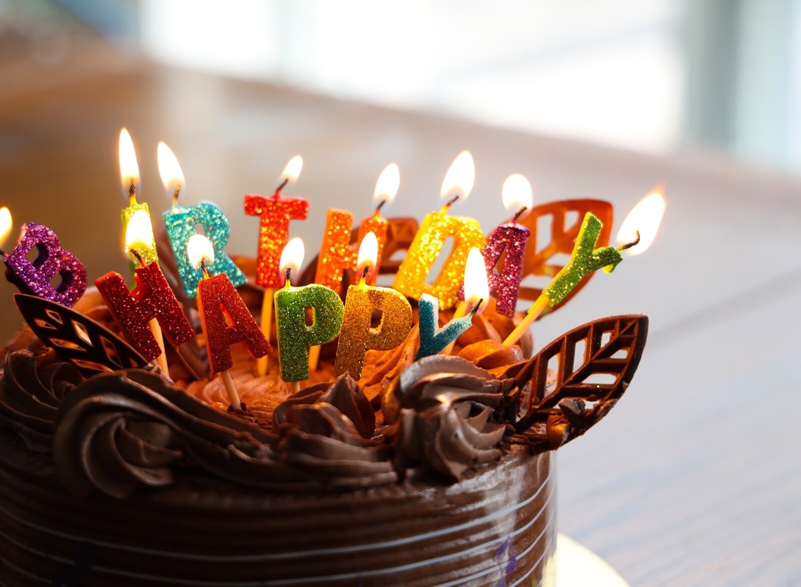 Zdjęcie przedstawia urodzinowy tort czekoladowy. Na torcie zostały ustawione palące się różnokolorowe świeczki urodzinowe, które układają się w napis w języku angielskim: Happy Birthday.