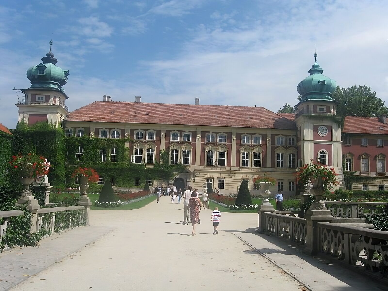Zdjęcie przedstawia zdjęcie zamku w Łańcucie od strony wejścia głównego, które prowadzi przez kamienny most nad dawną fosą. Kamienna balustrada mostu ozdobiona jest kamiennymi wazonami z kwiatami. Przez most aleją do wejścia podążają ludzie, w tym dzieci. W tyle zdjęcia jest widok obiektu głównego. To dwukondygnacyjny gmach z wysokim podpiwniczeniem ozdobionym boniowaniem. Po bokach budynku są ryzality w formie wież nakryte baniastymi hełmami. Pod nimi w każdej wieży jest balkon, a pod nim na wieży po prawej stronie zdjęcia jest umieszczony zegar. Po prawej stronie zdjęcia za wieżą widoczny jest dwukondygnacyjny fragment skrzydła zamku. Po lewej stronie obrazu widać na budynku pnącą się winorośl. Przed gmachem są rabaty kwiatowe i krzewy ogrodu francuskiego. 