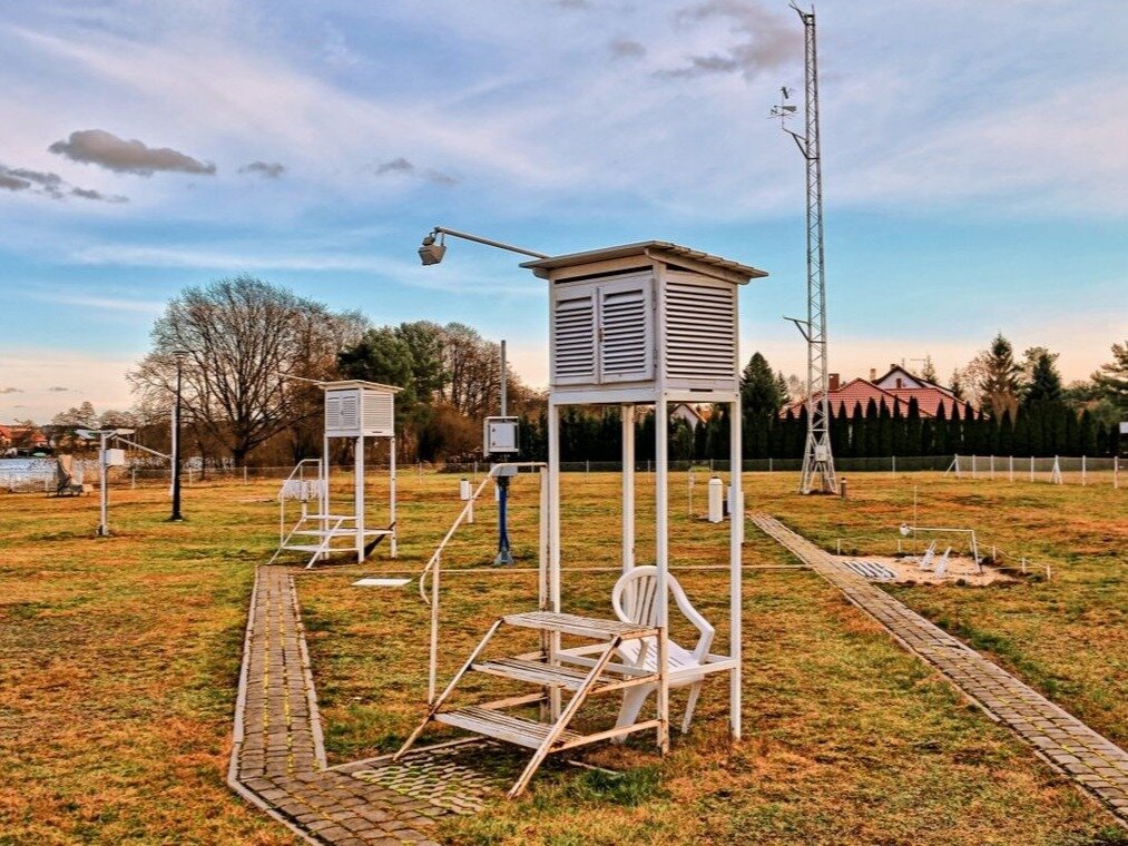 Fotografia przedstawiająca stację hydrologiczną. Na płaskim ogrodzonym terenie widać metalową konstrukcję z platformą na górze. Prowadzą na nią metalowe schody. W tle widać drzewa i stojące wśród nich budynki.