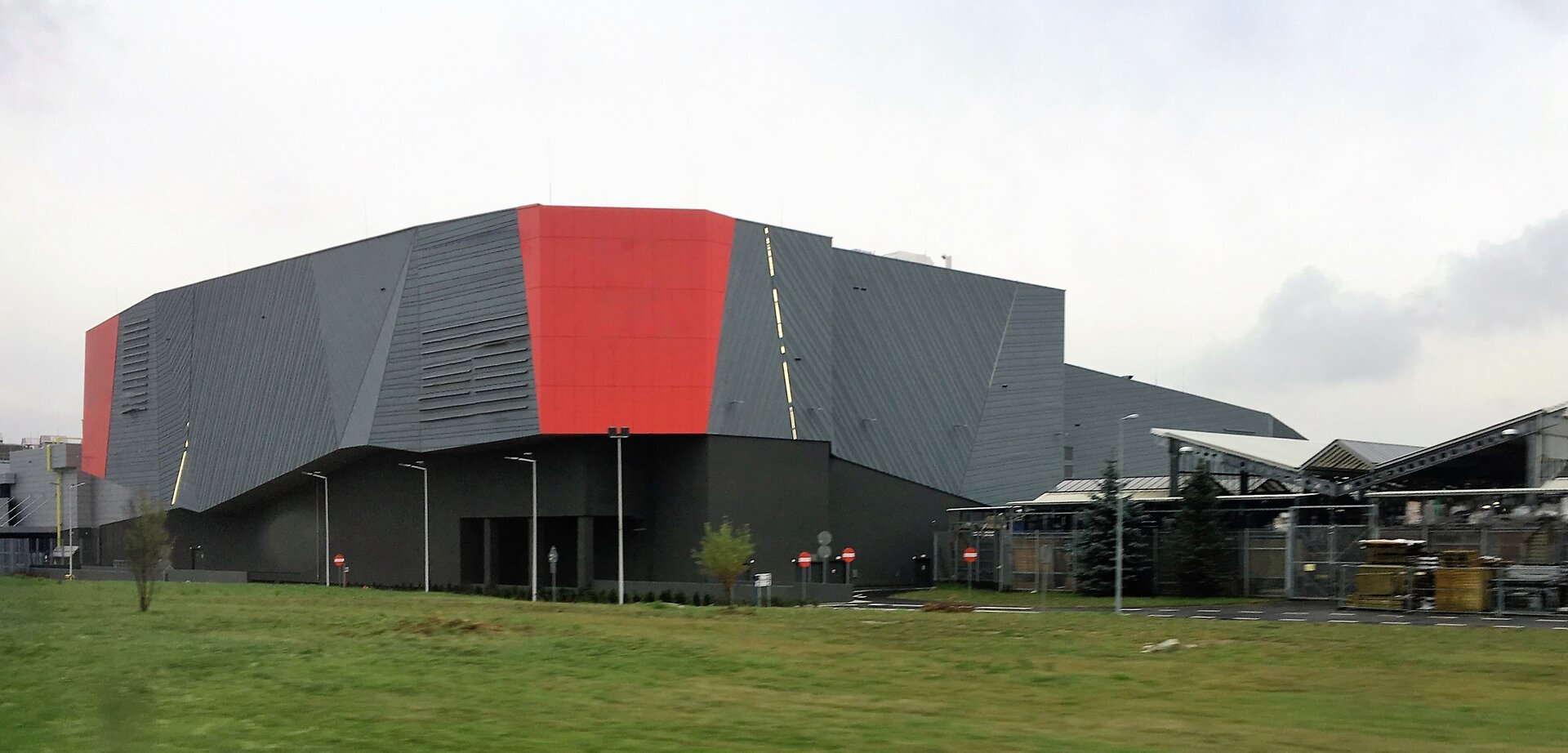 Na fotografii budynek o nowoczesnej architekturze, nieregularnej, poziomej bryle. Na fasadzie logotypy sklepów i innych usługodawców prowadzących działalność na terenie budynku. Na pierwszym planie trawa, płaski teren. U dołu bryły wjazd do budynku, brak okien w ścianach.