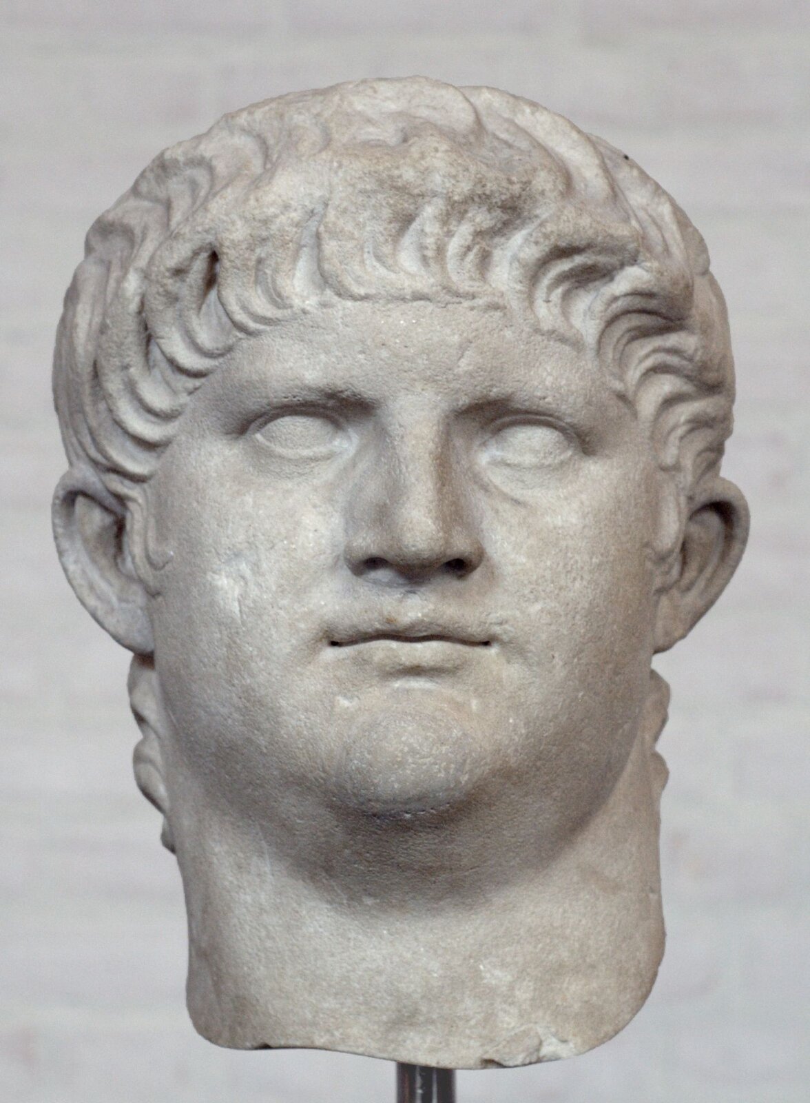 Ilustracja przedstawia rzeźbę głowy Nerona z białego marmuru. Jest to mężczyzna z krótkimi włosami opadającymi na czoło. Ma niewielkie zamknięte usta. Oczy nie mają wyrzeźbionych źrenic. Twarz jest okrągła.