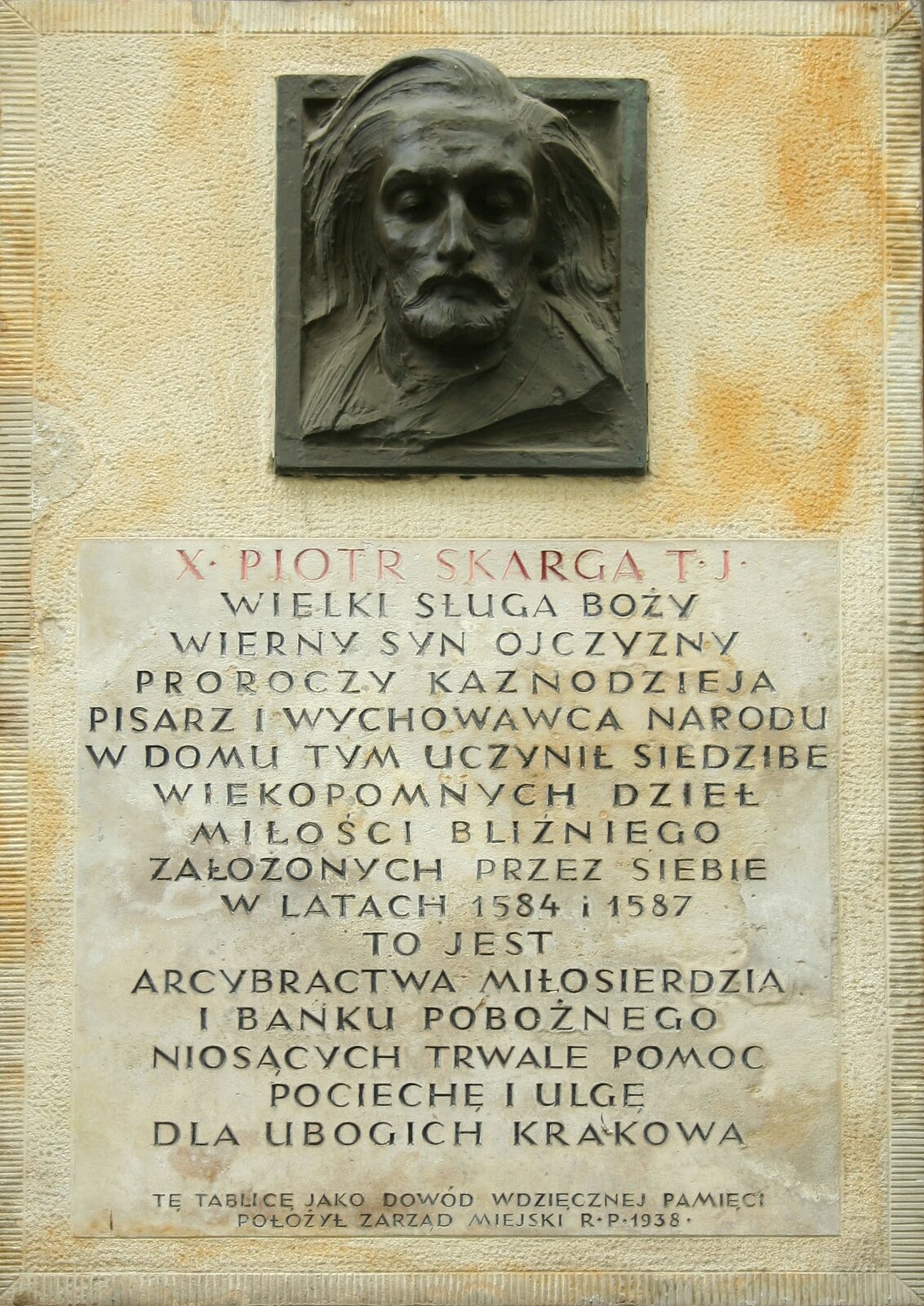 Ilustracja przedstawia pomnik w postaci wmurowanej płyty – płaskorzeźby. Na płaskorzeźbie widać twarz mężczyzny z długimi włosami do ramion, wąsami i brodą. Pod tablicą widnieje napis: X Piotr Skarga TJ Wieli Sługa Boży, Wierny Syn Ojczyzny, Proroczy Kaznodzieja, Pisarz i wychowawca narodu, w domu tym uczynił siedzibę wiekopomnych dzieł miłości bliźniego założonych przez siebie w latach 1584 i 1587 to jest arcybractwa miłosierdzia i banku pobożnego niosących trwale pomoc pociechę i ulgę dla ubogich z Krakowa. Tę tablicę jako dowód wdzięcznej pamięci położył Zarząd Miejski RR 1938