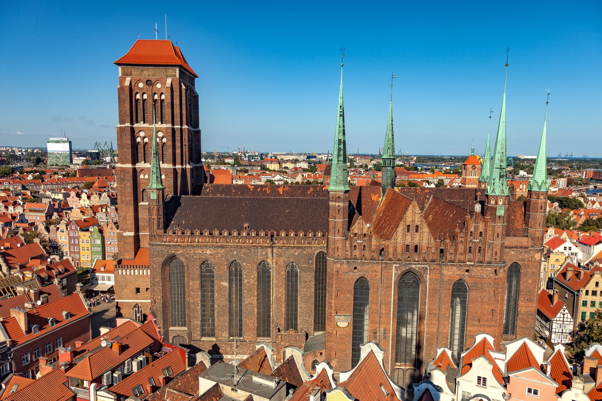 Zdjęcie Bazyliki Mariackiej w Gdańsku. Ukazuje obiekt zbudowany z cegły w stylu gotyckim z masywną wieżą dzwonną i smukłymi narożnymi wieżyczkami. W elewacji znajdują się wysokie, ostrołukowe okna o mocno rozglifionych ościeżach, ze skromną dekoracją maswerkową. Na szerokim transepcie znajdują się smukłe wieżyczki. W tle rozciąga się panorama Starego Miasta.