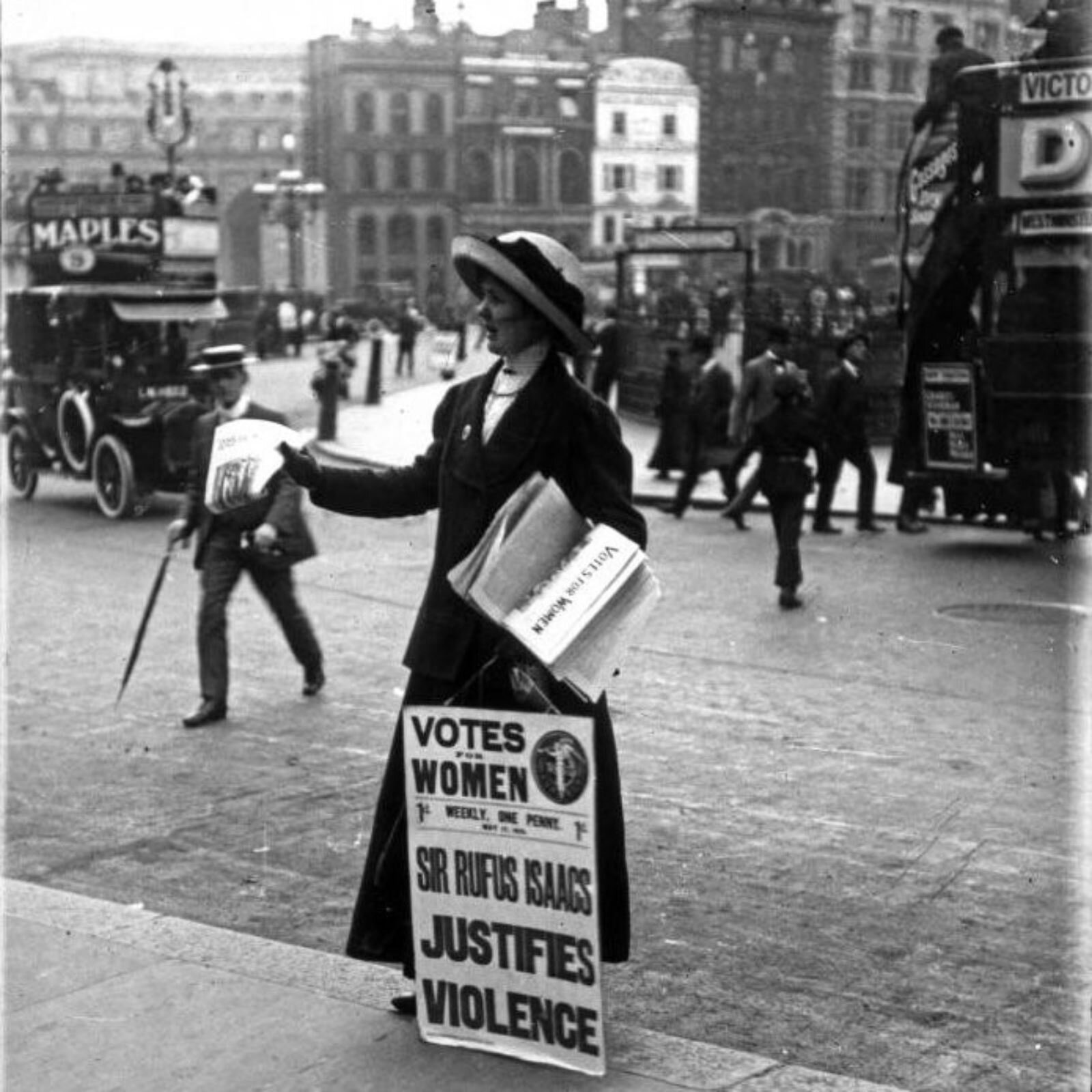Czarno‑białe zdjęcie przedstawia kobietę stojącą na chodniku i trzymającą transparent i gazety. Kobieta jest ubrana w długą ciemną spódnicę i żakiet, na głowie ma jasny kapelusz z czarną kokardą. Kobieta zwrócona jest w lewo, prawą rękę ma wyciągniętą do przodu i trzyma w niej gazetę, a lewą ręką przyciska do boku stos gazet. Przed nią widoczny jest transparent z napisem w języku angielskim: VOTES FOR WOMEN. WEEKLY ONE PENNY. SIR RUFUS ISAACS JUSTIFIES VIOLENCE. W tle widoczna jest ulica, przez którą przechodzą ludzie. Po prawej stronie ulicy stoją pojazdy, a w tle widoczne są budynki.