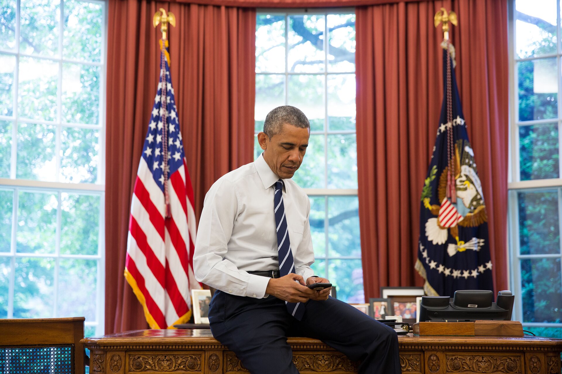 Zdjęcie przedstawia prezydenta USA, Baraka Obamę. Mężczyzna siedzi na biurku w Gabinecie Owalnym. Trzyma w rękach telefon komórkowy. W tle wykusz z dużymi oknami.