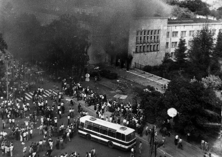 Czarno-biała fotografia przedstawiająca płonący budynek Wojewódzkiego Komitetu PZPR w Radomiu. Wokół tłum demonstrantów