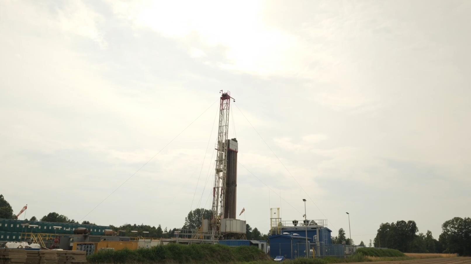 Na zdjęciu widać maszt urządzenia wiertniczego znajdującego się na placu wiertniczym.Widoczne na zdjęciu urządzenie wiertnicze jest w kształcie ostrosłupa ściętego. W dolnej części zdjęcia widoczny jest podest na którym znajdują się dwa białe kontenery. Pomiędzy kontenerami umieszczona jest wieża wiertnicza. Wieża jest konstrukcją kratową o kształcie cienkiego podłużnego prostokąta o orientacji pionowej. Z boku wieży odchodzą stalowe liny. Są to odciągi. Po bokach wieży zamontowane są długie rury. Jest to ich miejsce magazynowe. Na górze wieży widać walec po którym biegnie w dół stalowa lina. Jest to wielokrążek górny. Na końcu liny widoczne jest urządzenie do którego przyłączona jest rura wiertnicza. Całość urządzenia widoczna jest z boku.Z boku urządzenia widać niebieskie prostopadłościenne zbiorniki. Są to zbiorniki płuczkowe. Na górze zbiorników znajduje się podest zabezpieczony barierką. Po lewej stronie urządzenia wiertniczego znajdują się szare kontenery ustawione w szeregu. Kontenery są ustawione w formie jeden and drugim.W tle zdjęcia widać tereny leśne.