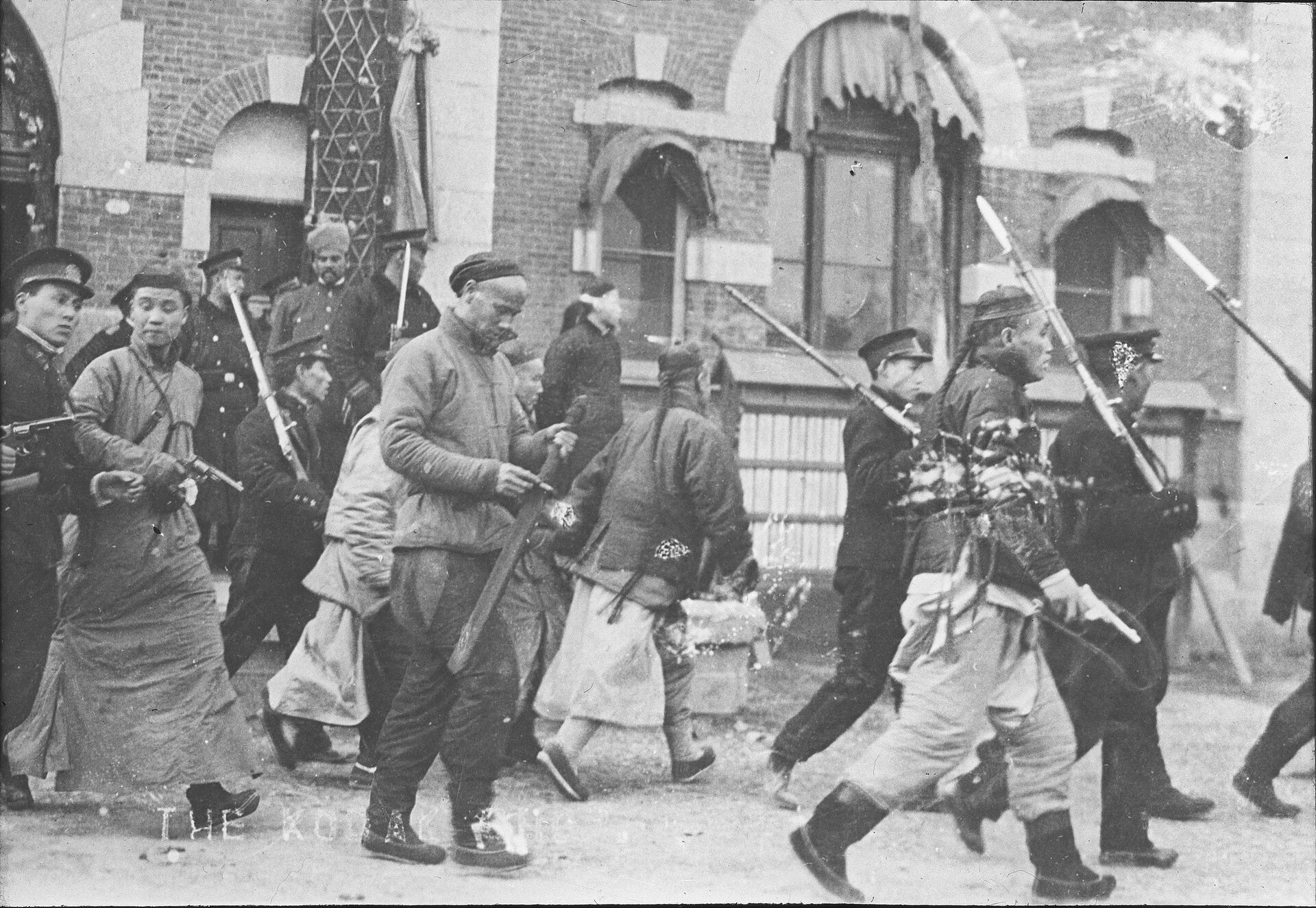 Zdjęcie przedstawia ludność chińską na jednej z ulic miasta w trakcie rewolucji w 1930 roku. Widzimy czterech mężczyzn ubranych w dawne chińskie stroje. Mają krótkie, watowane kurtki, szerokie spodnie zwężające się do dołu, niektórzy mają jeszcze długie spódnice, miękkie buty do pół łydki, a na ogolonych głowach okrągłe czapeczki. Jedynie z tyłu mają pozostawiony długi kosmyk włosów zapleciony w warkocz. Dwóch z nich ma pistolety, a jeden miecz. Pomiędzy nimi widzimy idących lub stojących żołnierzy. Ubrani są w kurtki, proste spodnie, okrągłe czapki z otokiem i daszkiem. Na ramionach trzymają karabiny z bagnetem. Kilku żołnierzy stoi przed wejściem do budynku, a jeden z nich ma nakrycie głowy przypominające turban. 