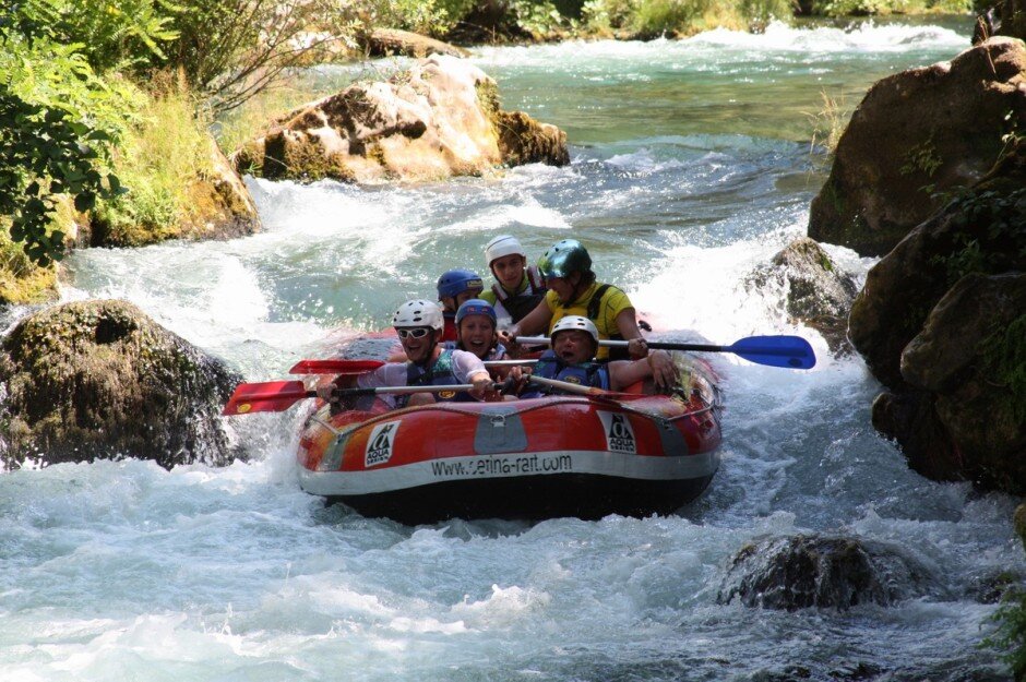 Na zdjęciu w czerwono czarnym pontonie znajduje się sześć osób płynących po wzburzonej wodzie. Po prawej i lewej stronie rzeki wąskiej rzeki widać skały i niewielkie krzewy, w tle część jeziora i skały, które tworzą kaskadę, po której spływa ponton. Ludzie na fotografii trzymają wiosła, mają na głowie kaski. Wszyscy ubrani są w kamizelki ratunkowe. Ich twarze są uśmiechnięte. 