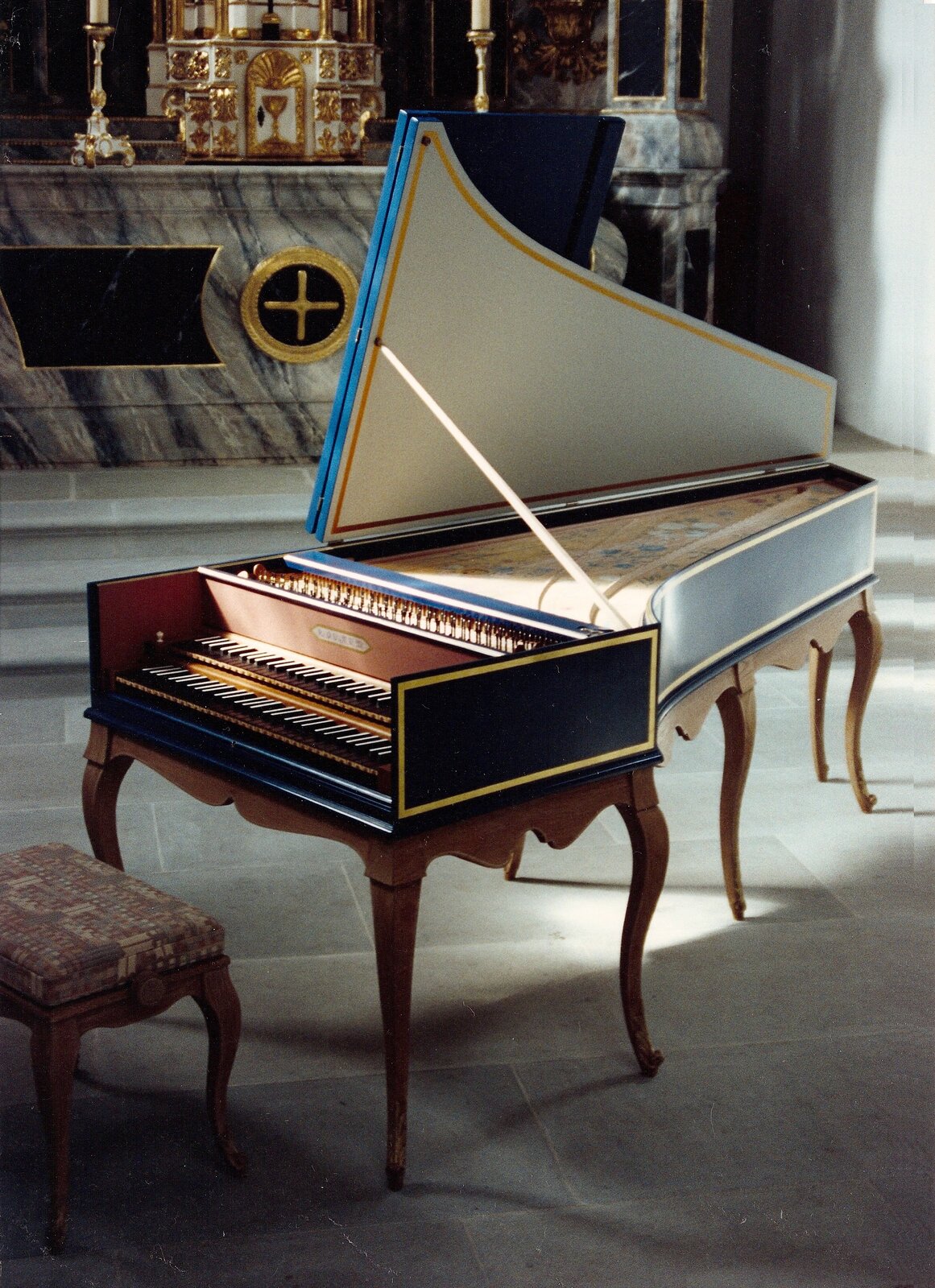 Fotografia przedstawia klawesyn. Instrument jest podobny do fortepianu, ale jest mniejszy. Ma podwójny ciąg klawiszy w dwóch kolorach, trójkątne pudło rezonansowe jest otwarte i przytrzymywane na cienkiej belce. Instrument jest drewniany, stoi na kilku drewnianych nogach. Przed nim znajduje się stołek z obitą materiałem poduszką. Całość stoi na podłodze z kamiennych płytek, w kościele, w tle znajduje się marmurowy ołtarz.