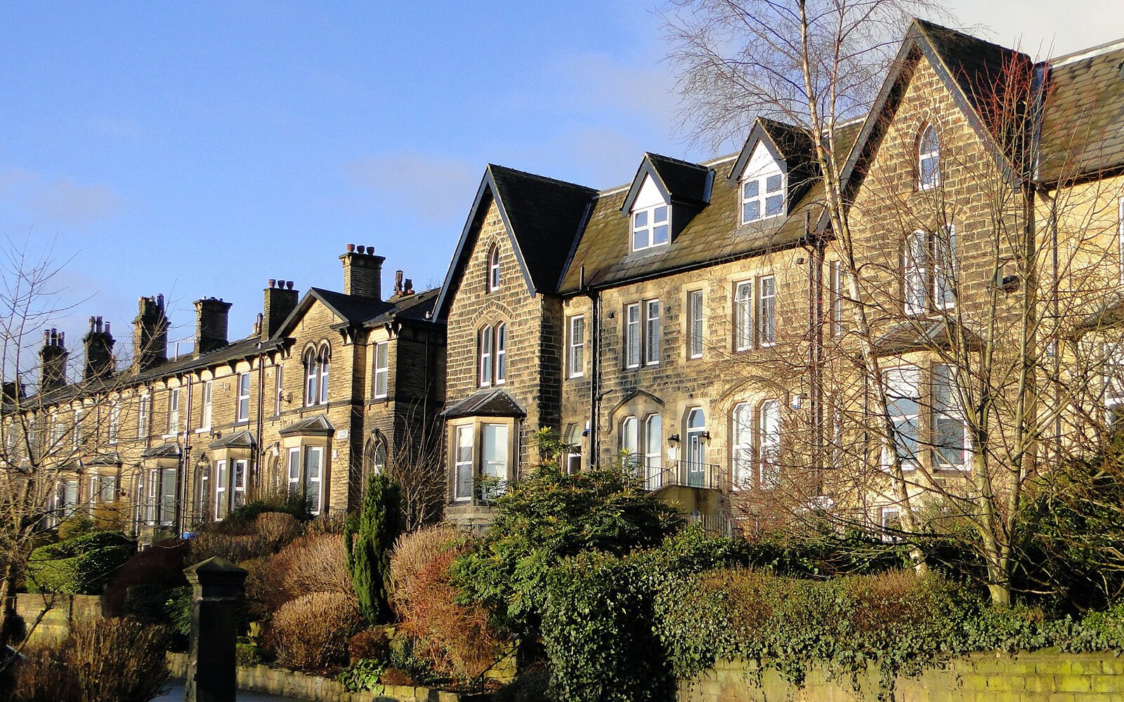 Zdjęcie przedstawia domy szeregowe. Na trzech z nich znajdują się kalenice, a reszta ma płaskie dachy na których są kominy. Domy mają po jednym oknie wykuszowym. Przed domami znajdują się przycięte krzewy. Przed krzewami znajduje się wąski kanał z wodą. Transkrypcja nagrań znajduje się poniżej.