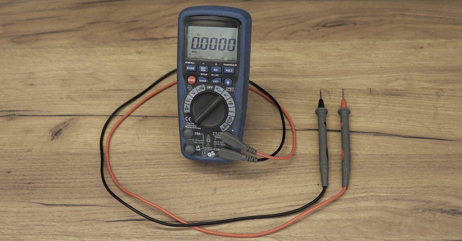 Zdjęcie przedstawia miernik wielkości elektrycznych. Składa się z niewielkiej jednostki centralnej z wyświetlaczem numerycznym, pokrętłem sterującym oraz przyciskami. Do gniazd urządzenia podłączone są za pomocą czerwonego i czarnego przewodu końcówki miernicze przypominające długopisy. Końcówki zakończone są metalowymi drucikami. 