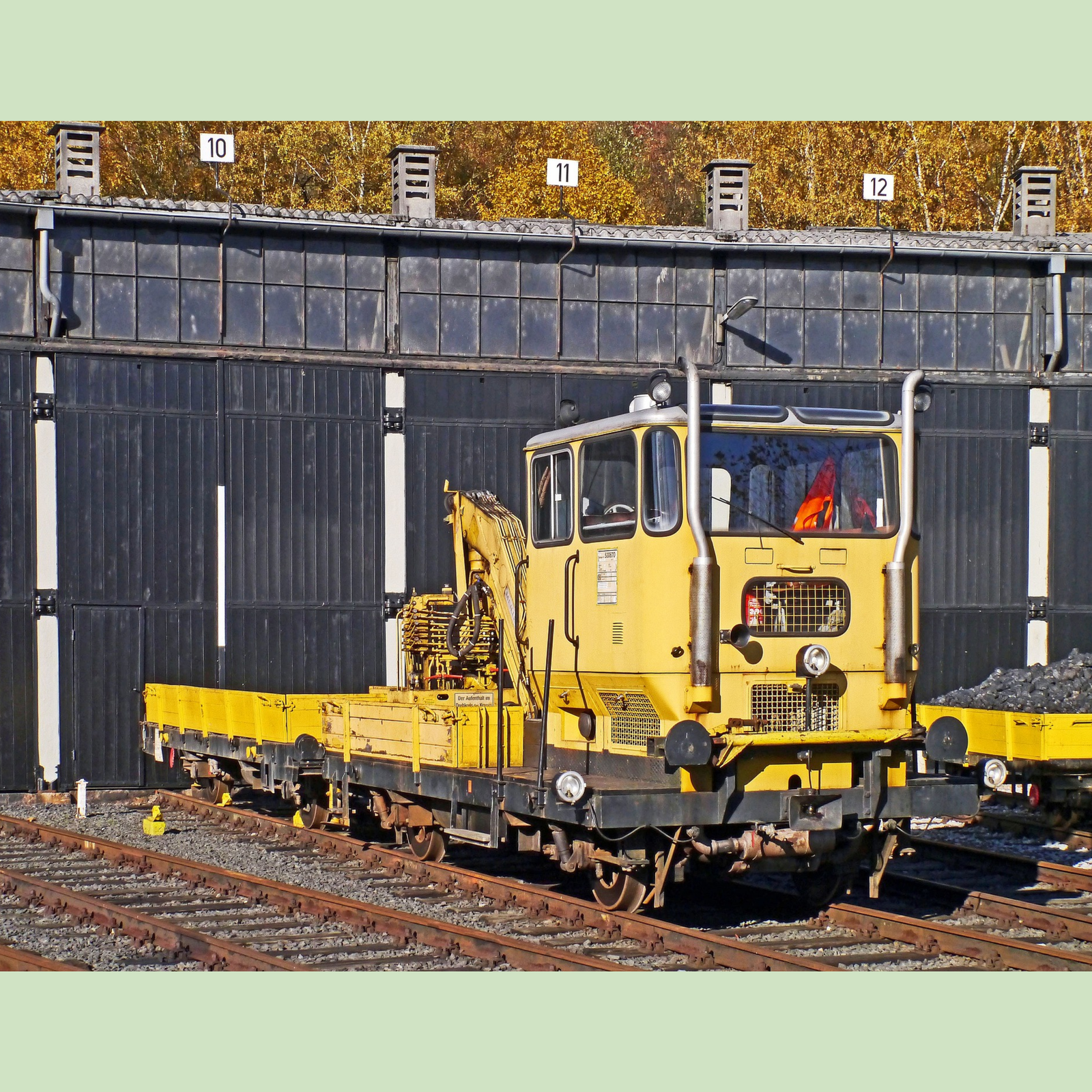 Zdjęcie przedstawia drezynę. Na przodzie płaskiej platformy znajduje się kabina, za którą jest skrzynia ładownicza i ramię żurawia.