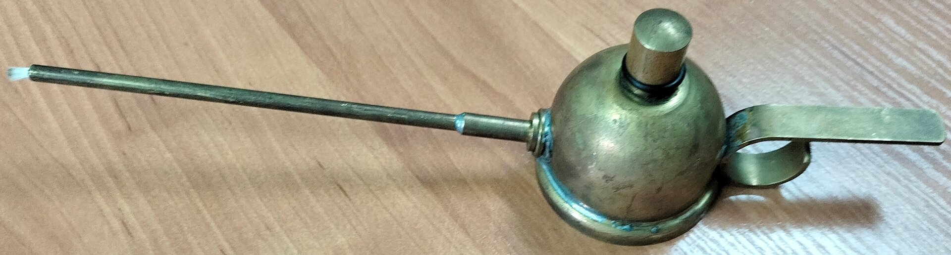 Zdjęcie przedstawia poglądowo lampkę spirytusową widzianą z góry. Widoczny jest półkolisty zbiornik z cylindrycznym pionowym elementem pośrodku. W lewo od zbiornika odchodzi cylindryczna rurka z wystającym na końcu białym knotem. W prawo od zbiornika odchodzi taśmowaty uchwyt.