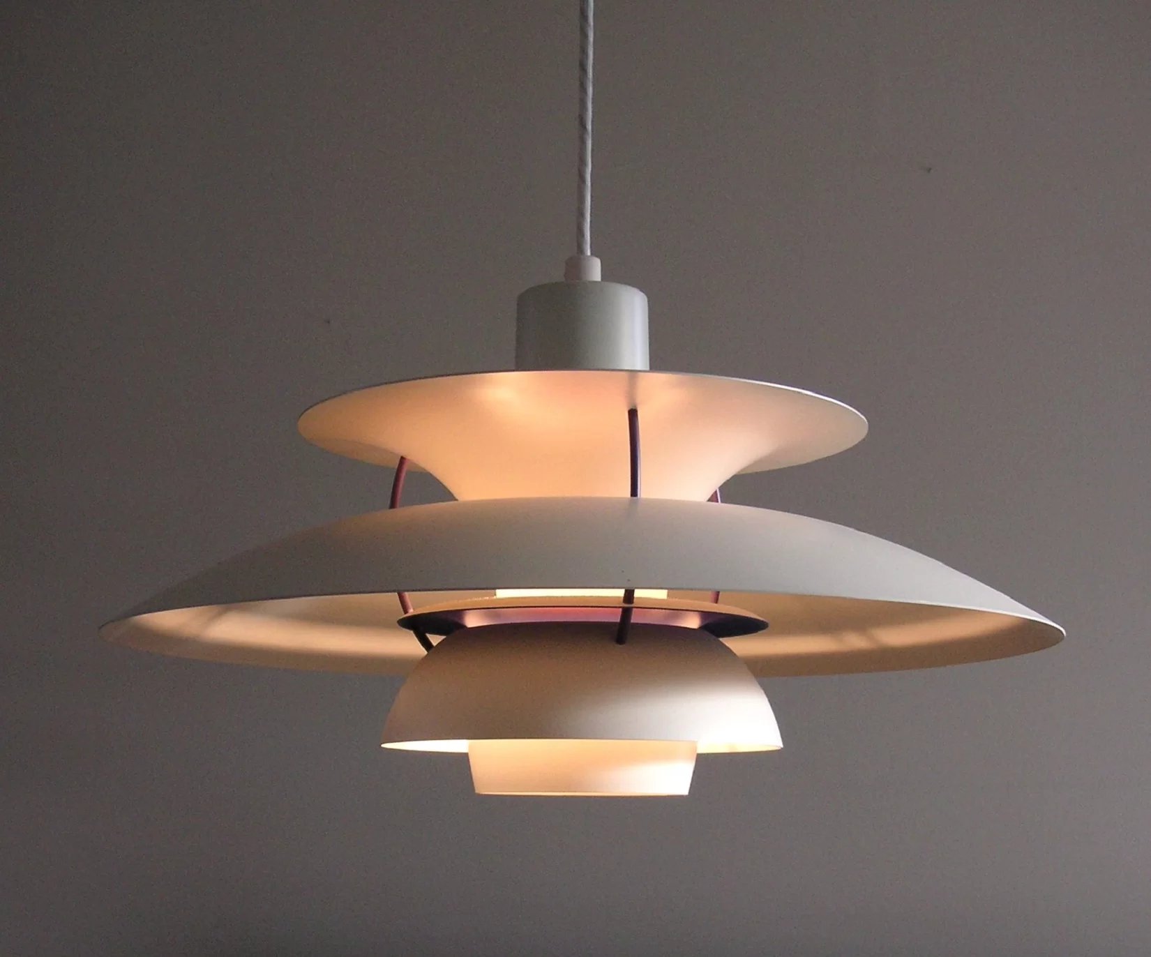Zdjęcie przedstawia lampę wiszącą na kablu na tle szarawej ściany. Lampa składa się z kilku zaokrąglonych kształtów w kolorze białym. Na samej górze, połączony z kablem jest kształt walca. Później element w kształcie lejka, w którym niżej umieszczono żarówkę. Pod nim znajduje się element w kształcie dużego, głębokiego talerza, ustawionego w dół. Następnie jeszcze jeden owalny kształt oraz element przypominający odwróconą w dół miskę. Z niego wystaje ostatni, owalny element. Ciepłe światło odbija się od poszczególnych elementów lampy.