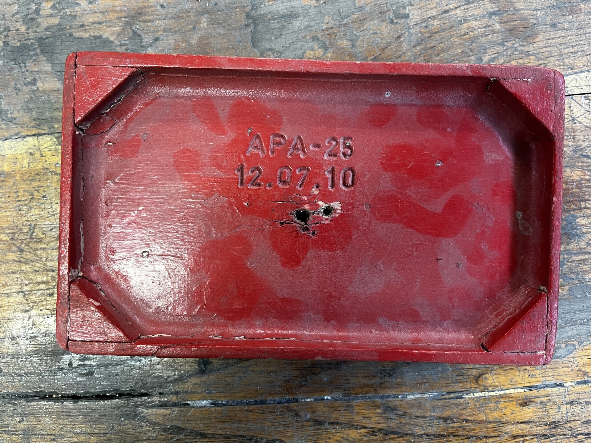 Na zdjęci pokazano drewniany model odlewniczy, który nie wymaga naprawy. Widoczny model jest w kształcie prostopadłościanu. Wnętrze prostopadłościany jest puste. Naroża są dodatkowo ścięte. W centralnej części modelu widać napis APA kreska dwadzieścia pięć. Niżej widoczna jest data dwunasty października dwa tysiące dziesięć. Pod datą znajdują się otwory przypominające przebicia. Są to otwory służące do wyciągania modelu z formy.