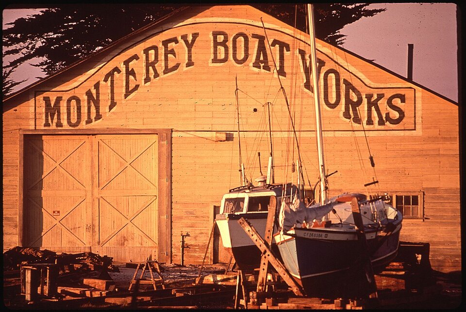 Ilustracja przedstawia upadły zakład budowy łodzi w Monterey. Jest to duży drewniany budynek z napisem Monterey Boat Works. Przed nim znajdują się dwie gotowe łodzie. Na ziemi porozrzucane są kawałki drewna oraz innych części. 