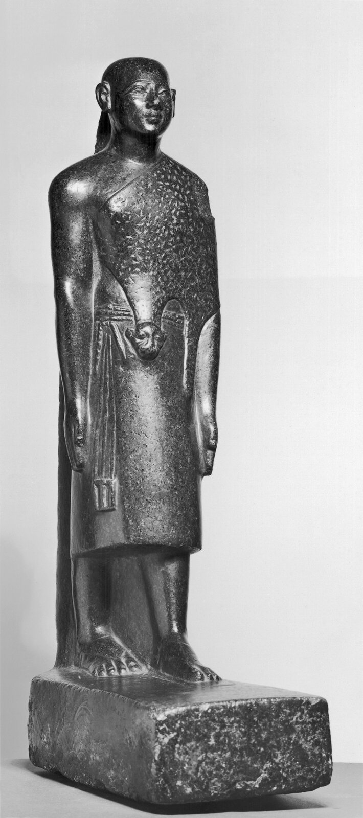 Na zdjęciu znajduje się czara figurka wyższego kapłana egipskiego. Nie ma on włosów i jest ubrany w szaty i zwierzęce skóry .