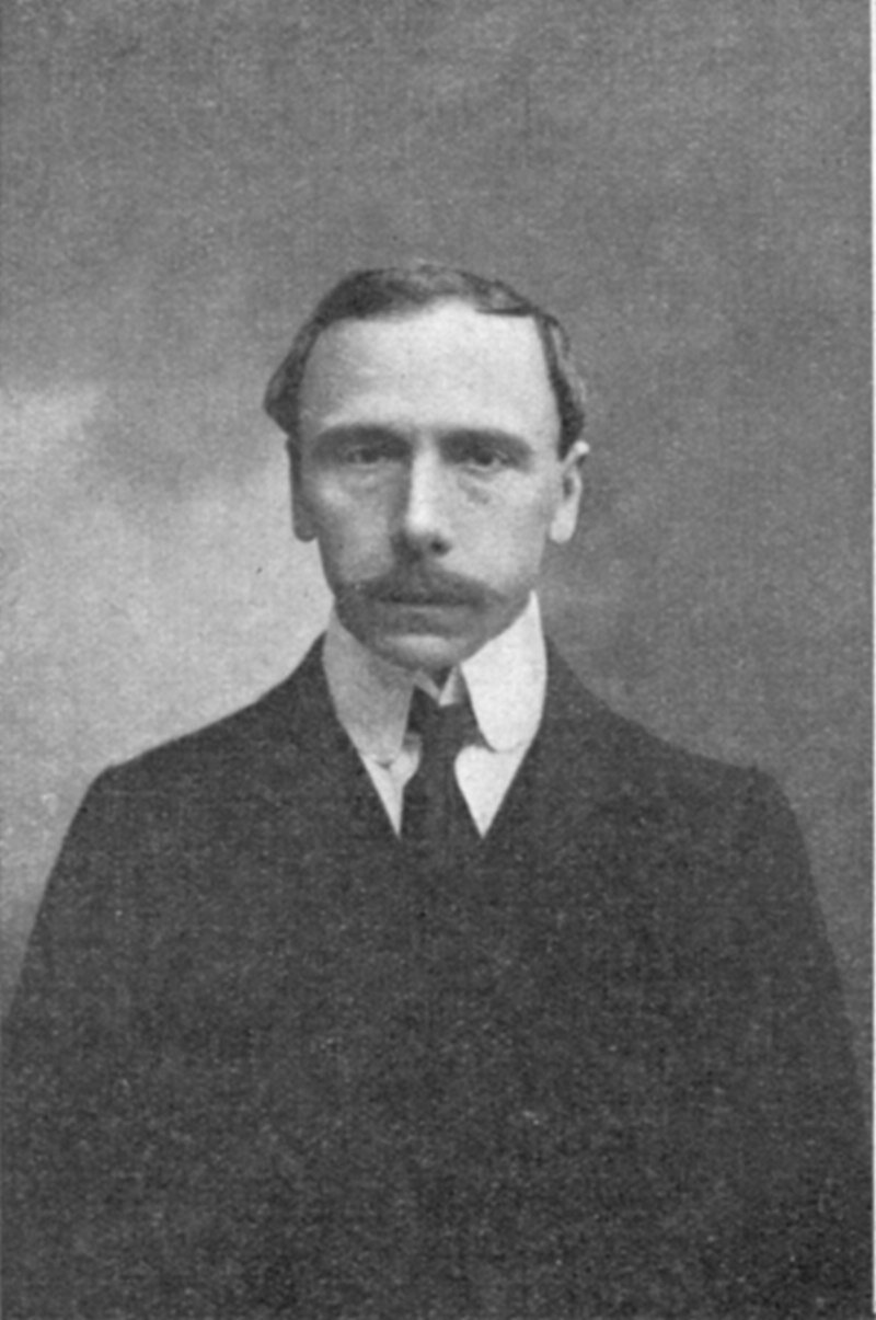 Zdjęcie przedstawia twarz dojrzałego mężczyzny. Nos mężczyzny jest duży, oczy głęboko osadzone, czoło wysokie, twarz szczupła. Mężczyzna ma krótkie, proste włosy. Ubrany jest w garnitur, koszulę i krawat.