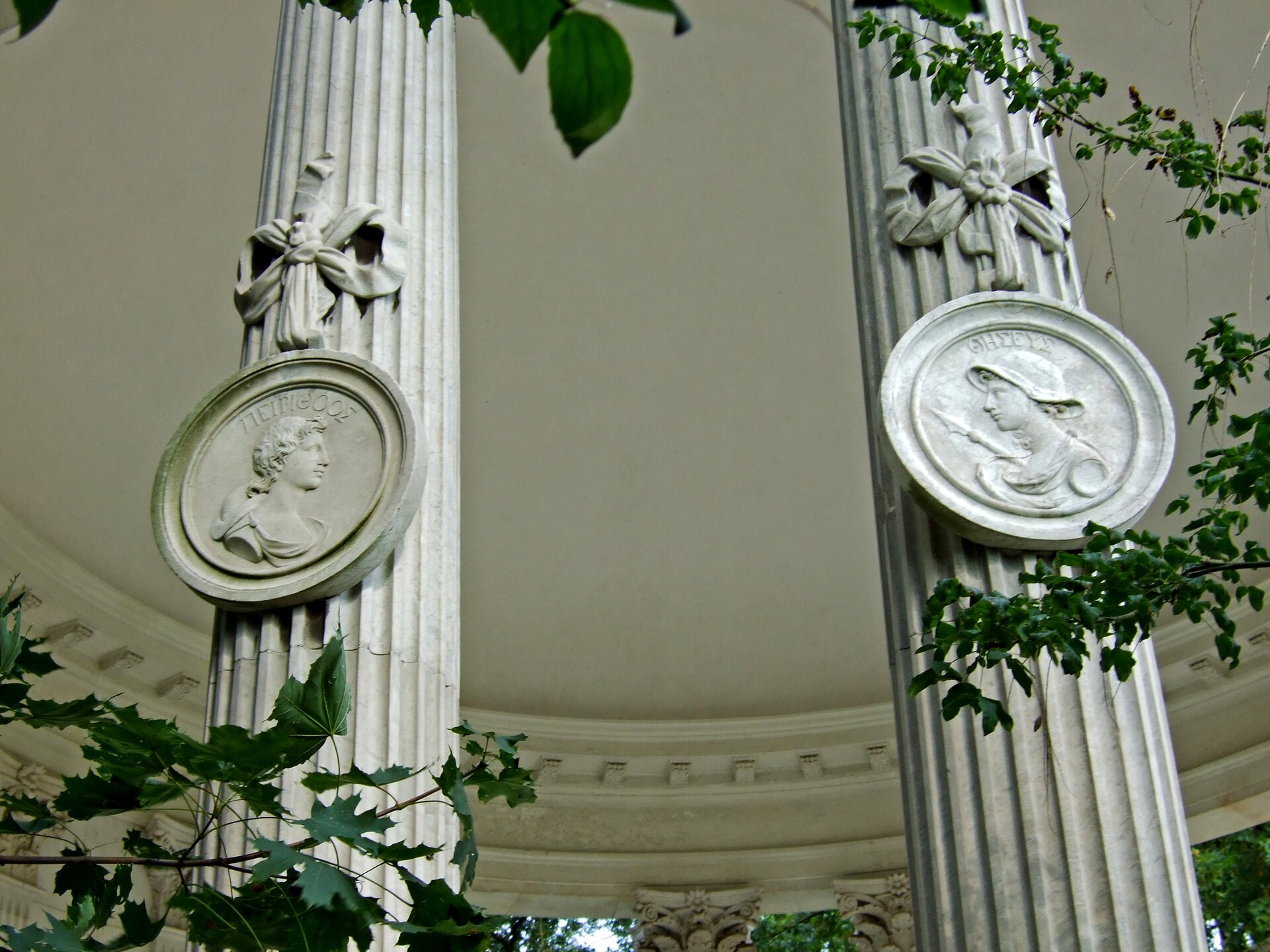 Fotografia przedstawia dwie kolumny z okrągłymi medalionami. Widoczne są na nich wizerunki mężczyzn z profilu. Mężczyzna na medalionie po lewej stronie ma kręcone włosy. Mężczyzna na medalionie z prawej strony ma na głowie hełm. Na głowami mężczyzn znajdują się napisy w języku greckim, są to ich imiona. Nad medalionami wyrzeźbiono duże kokardy. Wokół kolumn widoczne są gałęzie drzew z liśćmi.