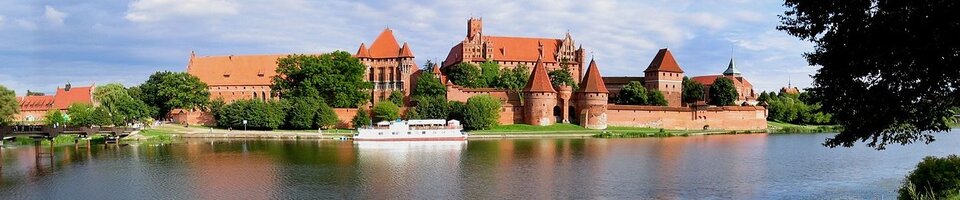 Na zdjęciu znajduje się zamek w Malborku. Jest on otoczony szeroką fosą. Prowadzi do niego drewniany most rozwieszony nad fosą. Zamek to potężna budowla posiadająca grube mury, ogromne baszty i wysokie budynki za murem. Przed murami rosną drzewa. 