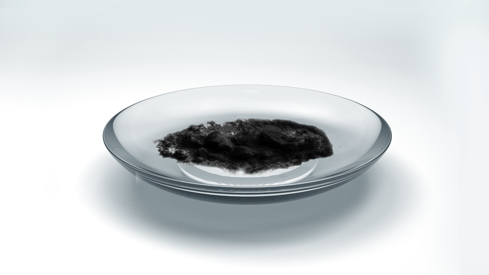 Zdjęcie przedstawia bromek żelaza(trzy). Czarny proszek znajduje się na szkiełku zegarkowym.