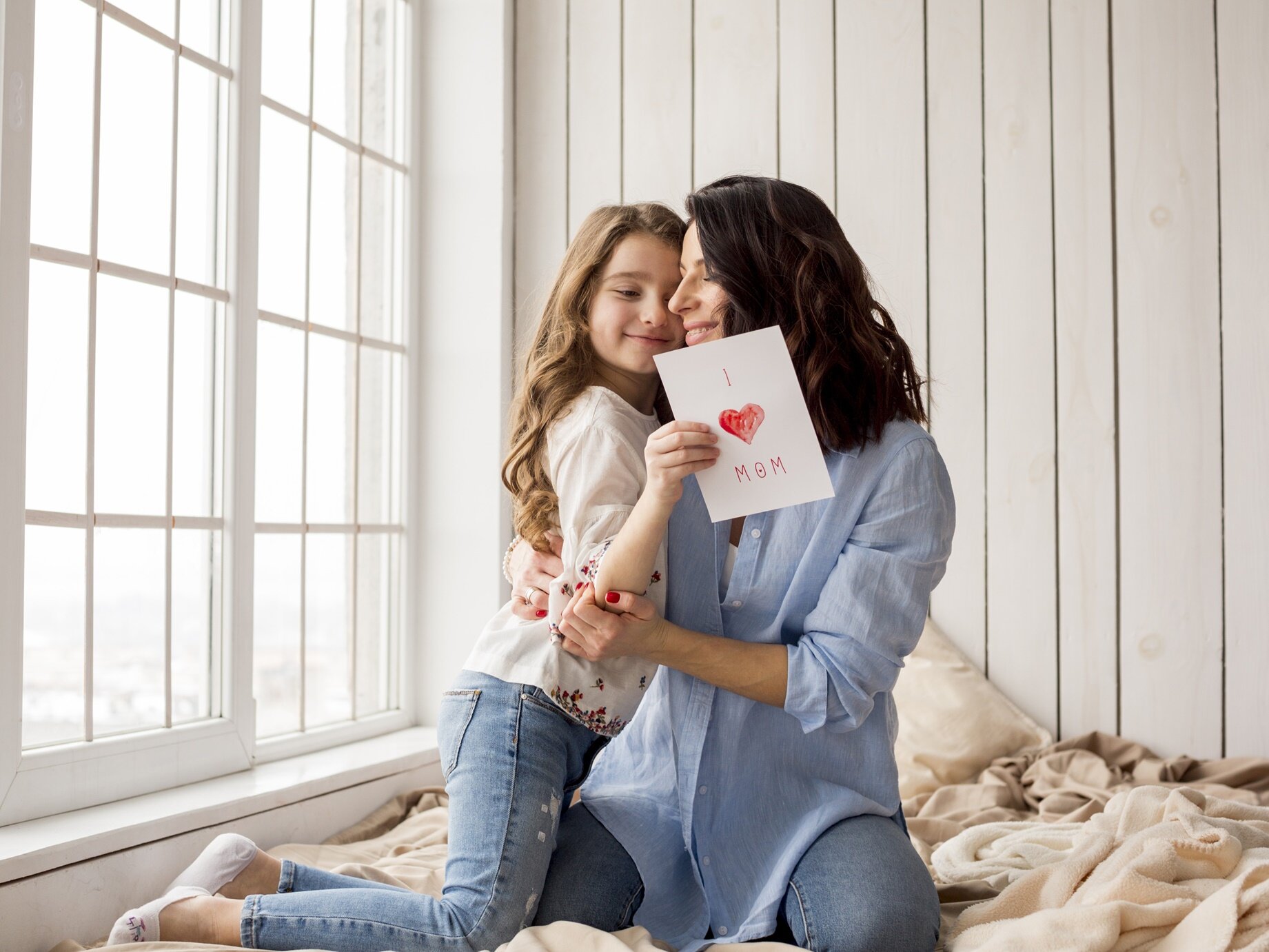 Zdjęcie przedstawia kobietę w średnim wieku przytulającą dziewczynkę w wieku szkolnym. Obie siedzą na łóżku. Dziewczynka trzyma w dłoniach kartkę z czerwonym sercem dla mamy. 