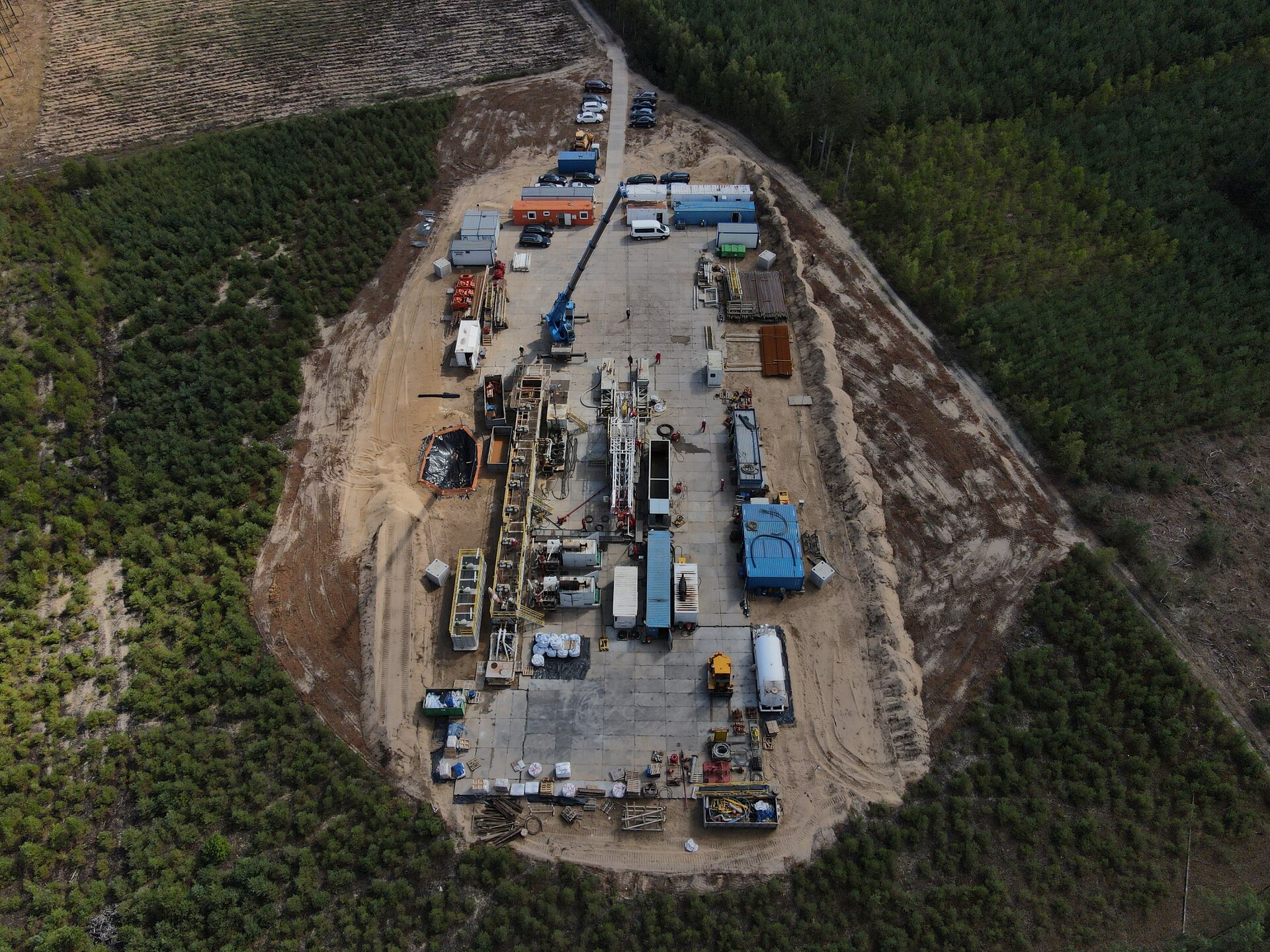 Na zdjęciu widać plac wiertniczy. Plac wiertniczy widoczny jest z lotu ptaka. Ma kształt prostokąta. Podstawa placu jest wyłożona betonowymi płytami. W centralnym punkcie placu znajduje się maszt wiertniczy. Maszt jest w kształcie ostrosłupa ściętego. Po lewej stronie masztu widać długi prostopadłościenny zbiornik. Na zbiorniku znajduje się podest zabezpieczony barierką. Jest to zbiornik płuczkowy. Po prawej stronie masztu widoczne są mniejsze prostopadłościenne zbiorniki. W prawym górnym rogu placu znajdują się kontenery pełniące rolę budynków biurowych.W lewym górnym rogu placu znajduje się żuraw na podwoziu samochodowym.W górnej części zdjęcia za terenem placu widoczny jest parking samochodowy. W tle zdjęcia widać tereny leśne.