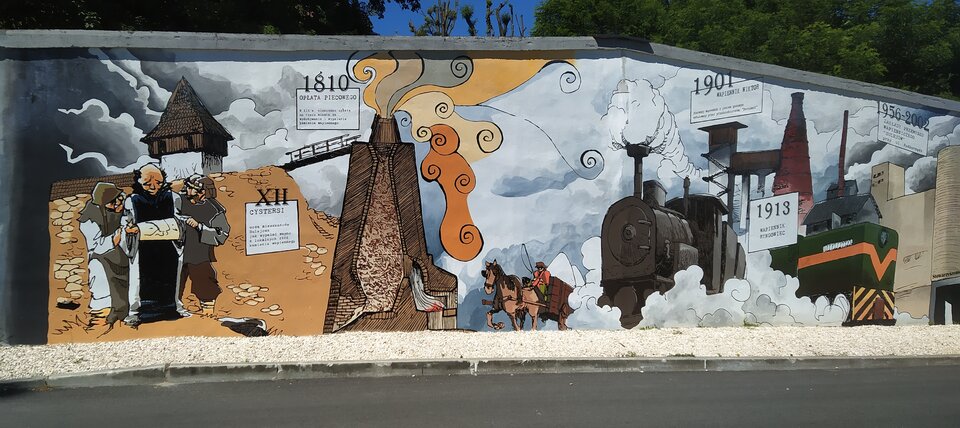 Fotografia ukazuje mural znajdujący się na murze przy ścieżce wyłożonej jasnymi kamieniami. Na muralu zaprezentowana została historię przemysłu wapienniczego. Od lewej strony: XII wiek - zakonnik trzymający mapę eskortowany jest w dwóch mężczyzn ubranych w ubrania robocze. W tle wieża wartownicza oraz most. 1810. Buchający ogniem piec, do którego zmierza woźnica siedzący na wozie, który ciągnie brązowy koń. 1901 - Wyoski komin do którego prowadzą schody. Komin zbudowany z czerwonej cegły. 1913 - Lokomotywa Parowa stoi na stacji - z komina i spod kół buchają kłęby pary. 1956‑2002 Nowoczesna fabryka - Zakłady Przemysłu Wapieniarskiego „Sulejów”. Przy fabryce elektrowóz.
