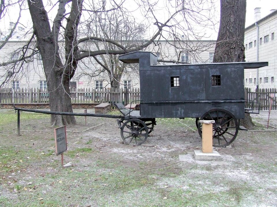 Zdjęcie przedstawia drewniany, kryty, czterokołowy wóz zwany kibitką. W wozie po obu stronach znajdują się dwa otwory okienne z kratami. Obok rosną drzewa, w tle widoczny jest budynek. 