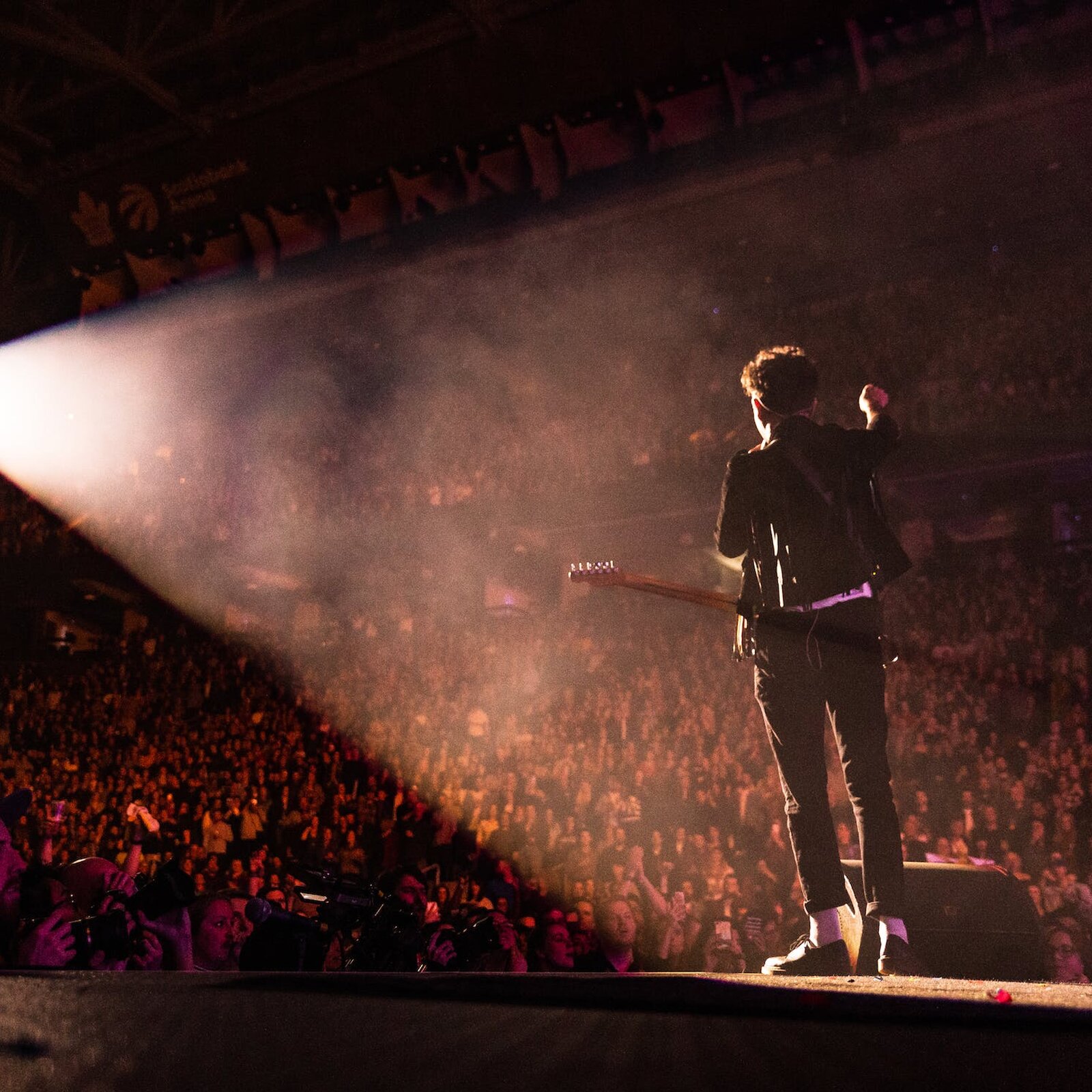Zdjęcie przedstawia osobę stojącą na scenie z gitarą. W dole widoczny tłum ludzi podnoszących ręce i robiących zdjęcia. 