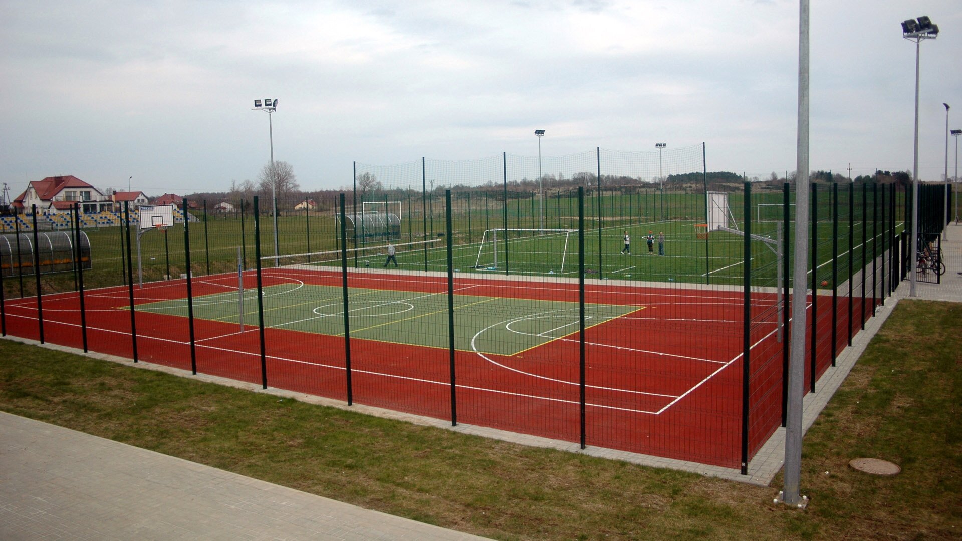 Fotografia przedstawiająca boiska do gry w koszykówkę, siatkówkę i piłkę nożną. Boiska otoczone są siatką. Na jednym z nich uczniowie graja w piłkę nożną.